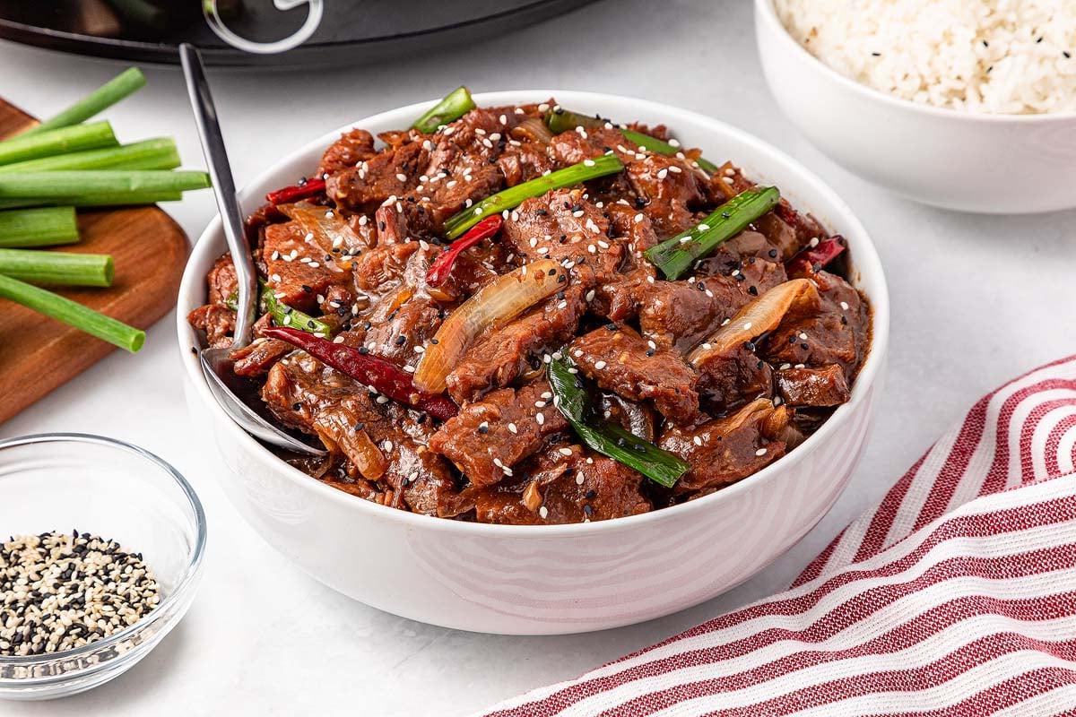 A bowl of slow cooker Mongolian beef garnished with green onions and sesame seeds sits enticingly on the table, accompanied by a bowl of rice, extra chopped green onions, and a dish of sesame seeds.