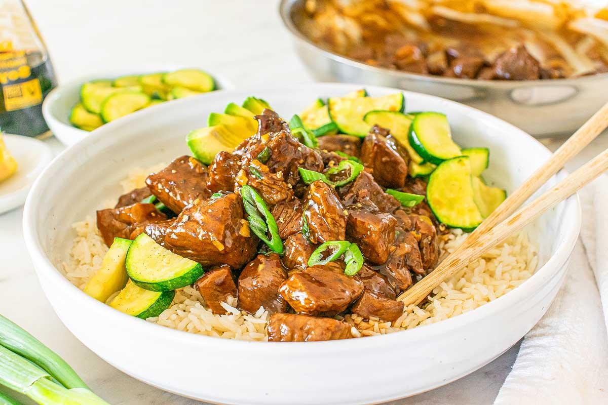 Μια μπολ stir-fry τεριγιάκι μοσχαριού με κολοκυθάκια σερβιρισμένο πάνω σε ρύζι, γαρνιρισμένο με φρέσκα κρεμμυδάκια, βρίσκεται φιλόξενα πάνω σε ένα τραπέζι δίπλα σε ένα τηγάνι και ένα μικρό πιάτο με κολοκυθάκια σε φέτες.