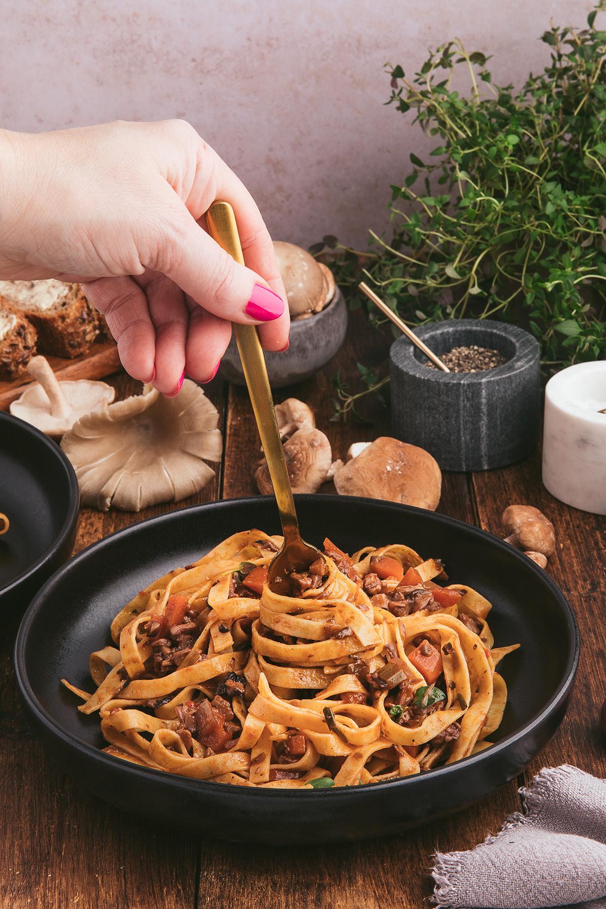 Ένα χέρι περιστρέφει fettuccine ζυμαρικά με Ραγού Μανιταριών και φρέσκα βότανα σε ένα μαύρο μπολ πάνω σε ένα ρουστίκ ξύλινο τραπέζι.