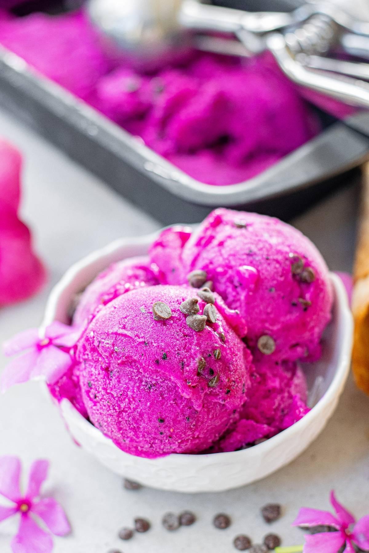 Φωτεινό ροζ παγωτό Dragon Fruit φτιαγμένο με μπανάνες και κομματάκια σοκολάτας σερβίρεται σε ένα λευκό μπολ, γαρνιρισμένο με ροζ λουλούδια για μια ζωντανή, πολύχρωμη γεύση παγωτού.