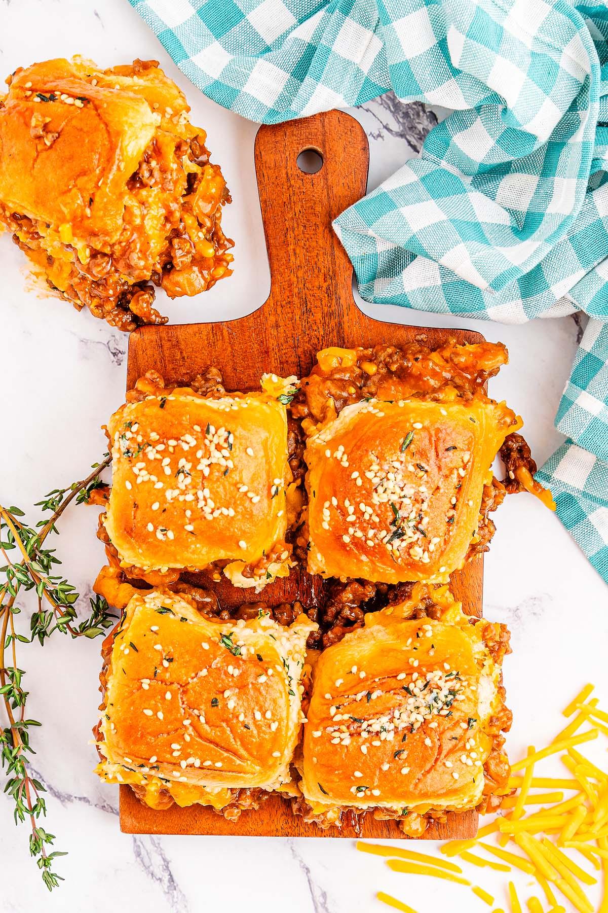 Τέσσερα ψημένα Sloppy Joe sliders με μοσχάρι και τυρί σε ψωμάκια με σουσάμι, παρουσιασμένα σε ξύλινη σανίδα και γαρνιρισμένα με μυρωδικά. Τριμμένο τυρί και μια καρό πετσέτα βρίσκονται κοντά.