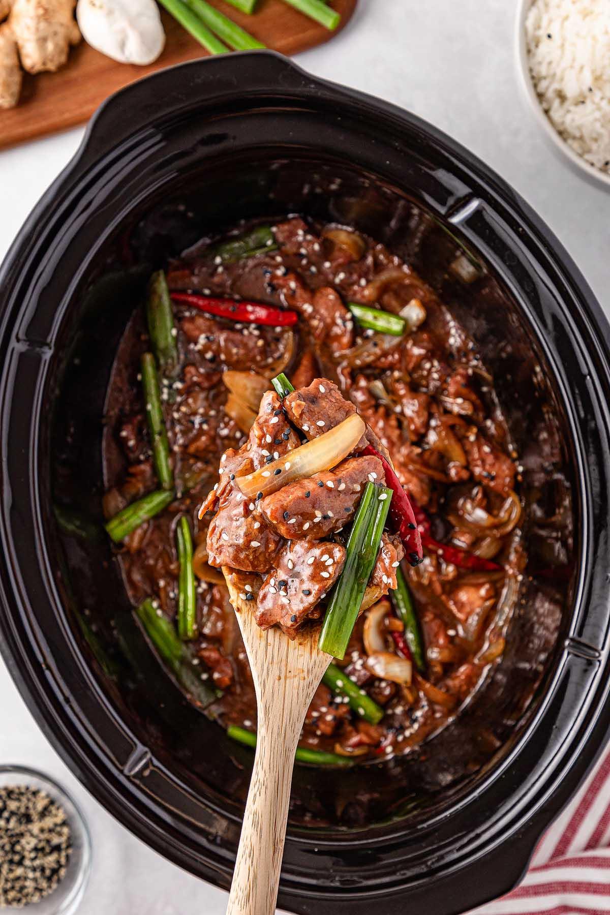 Slow cooker Mongolian beef stir-fry with sliced beef, fresh vegetables, and sesame seeds, being served with a wooden spoon.