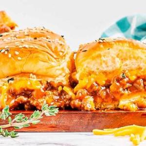 Κοντινό πλάνο ζουμερών Sloppy Joe sliders με λιωμένο τυρί και κιμά, με σουσάμι σε ψωμάκια, γαρνιρισμένα με φρέσκο θυμάρι, και ένα καρό ύφασμα στο φόντο.