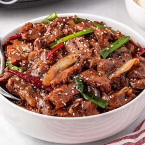 A bowl of stir-fried slow cooker Mongolian beef with green onions and sesame seeds rests on a white surface, accompanied by rice and chopped green onions.