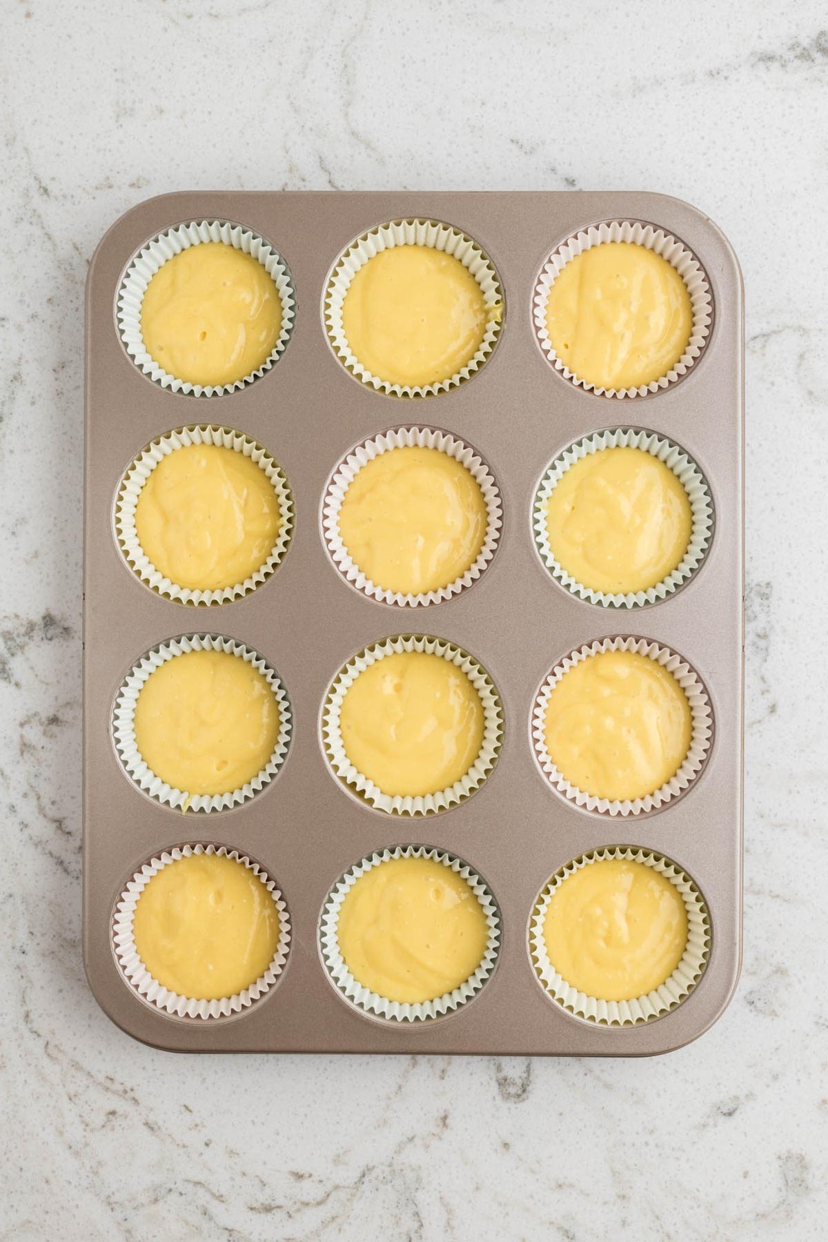 Ταψί για muffins γεμάτο με ωμό κίτρινο μείγμα σε δώδεκα χάρτινες θήκες, τοποθετημένο σε μαρμάρινο πάγκο.