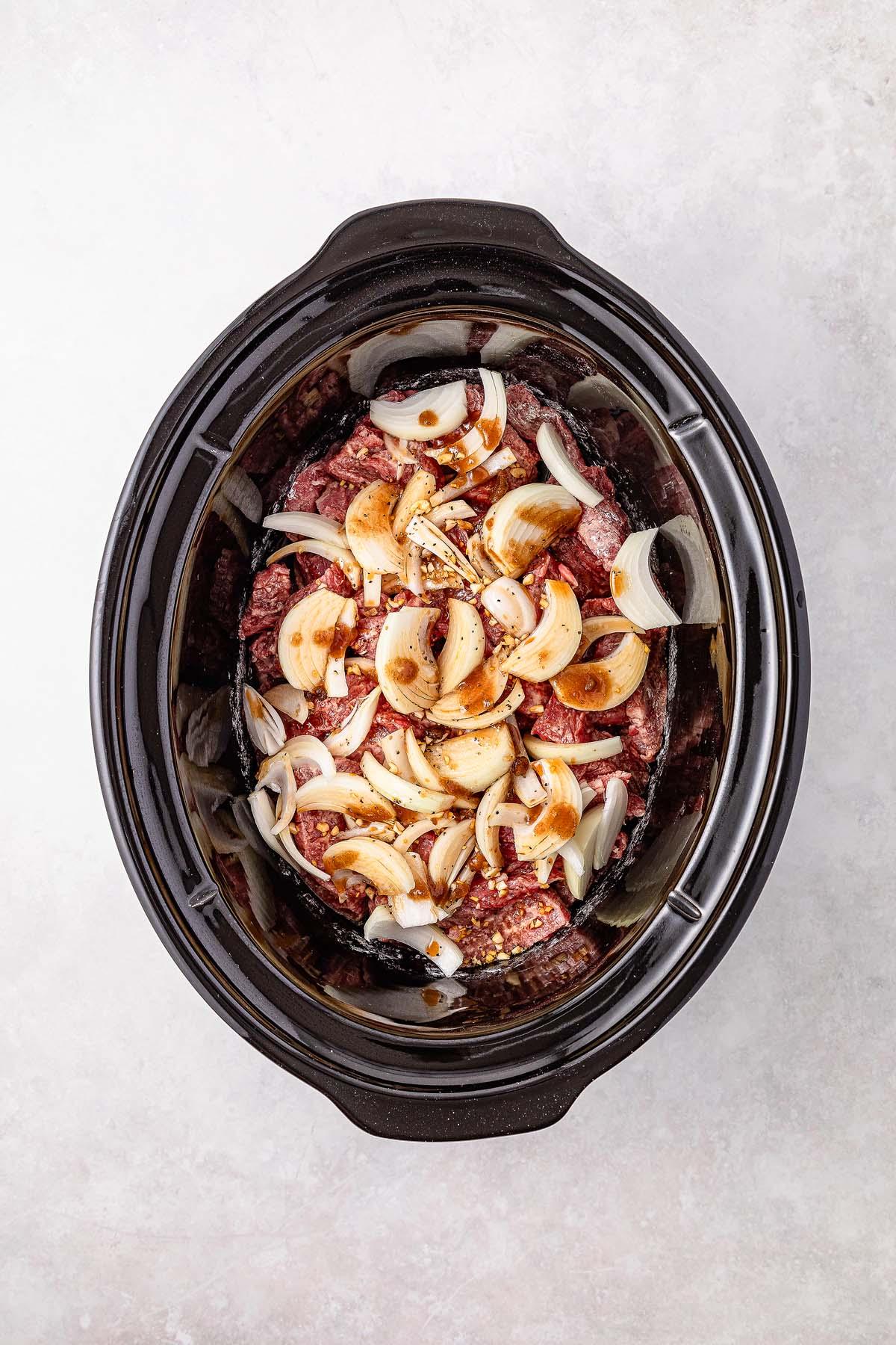 Savor the simplicity of slow cooker Mongolian beef with raw beef slices, chopped onions, and spices gently mingling on a pristine white surface.