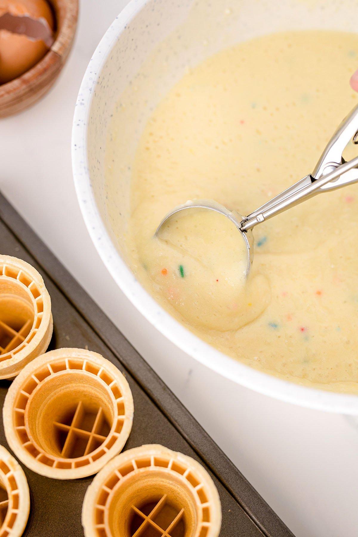 Ένα μπολ με μείγμα κέικ funfetti με μια μεζούρα βρίσκεται δίπλα σε μια φόρμα για muffins γεμάτη με cupcakes σε χωνάκια παγωτού, τέλεια για πάρτι γενεθλίων.