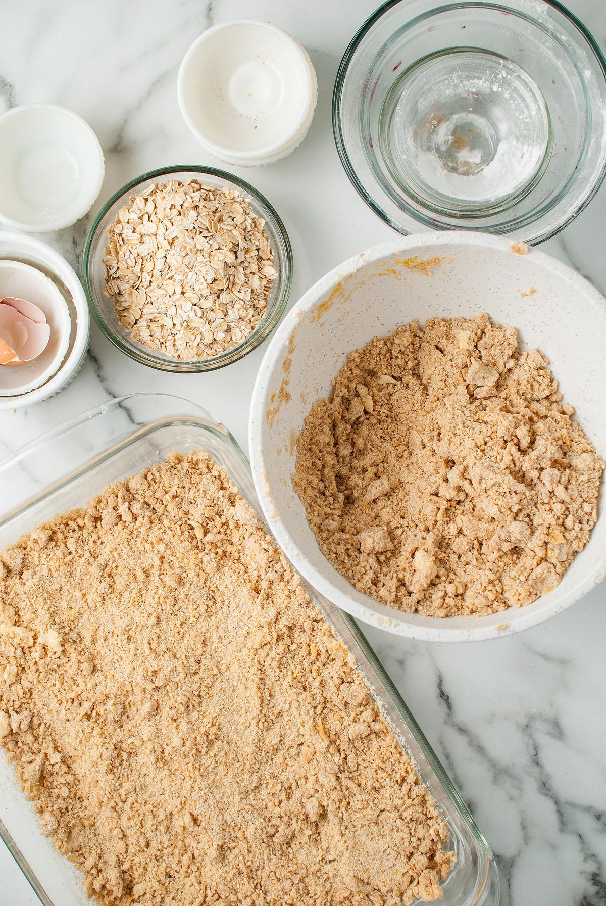 Γυάλινο ταψί με επικάλυψη crumble, μπολ βρώμης, μείγμα crumble, τσόφλια αυγών και γυάλινα μπολ σε μάρμαρο—ιδανικά για την παρασκευή μπαρών βρώμης με κεράσι Michigan τύπου Starbucks ή των αγαπημένων σας μπαρών κερασόπιτας.