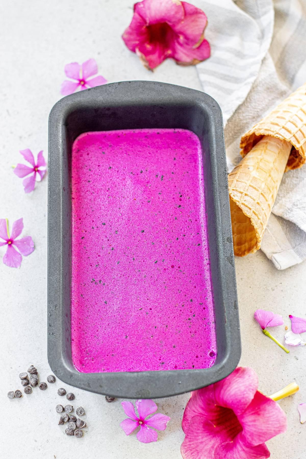 Ένα ταψί με φωτεινό ροζ παγωτό Dragon Fruit—ένα πολύχρωμο παγωτό—δίπλα σε χωνάκια, ροζ λουλούδια και διάσπαρτα κομματάκια σοκολάτας για μια ζωντανή εμφάνιση παγωτού χωρίς γαλακτοκομικά.