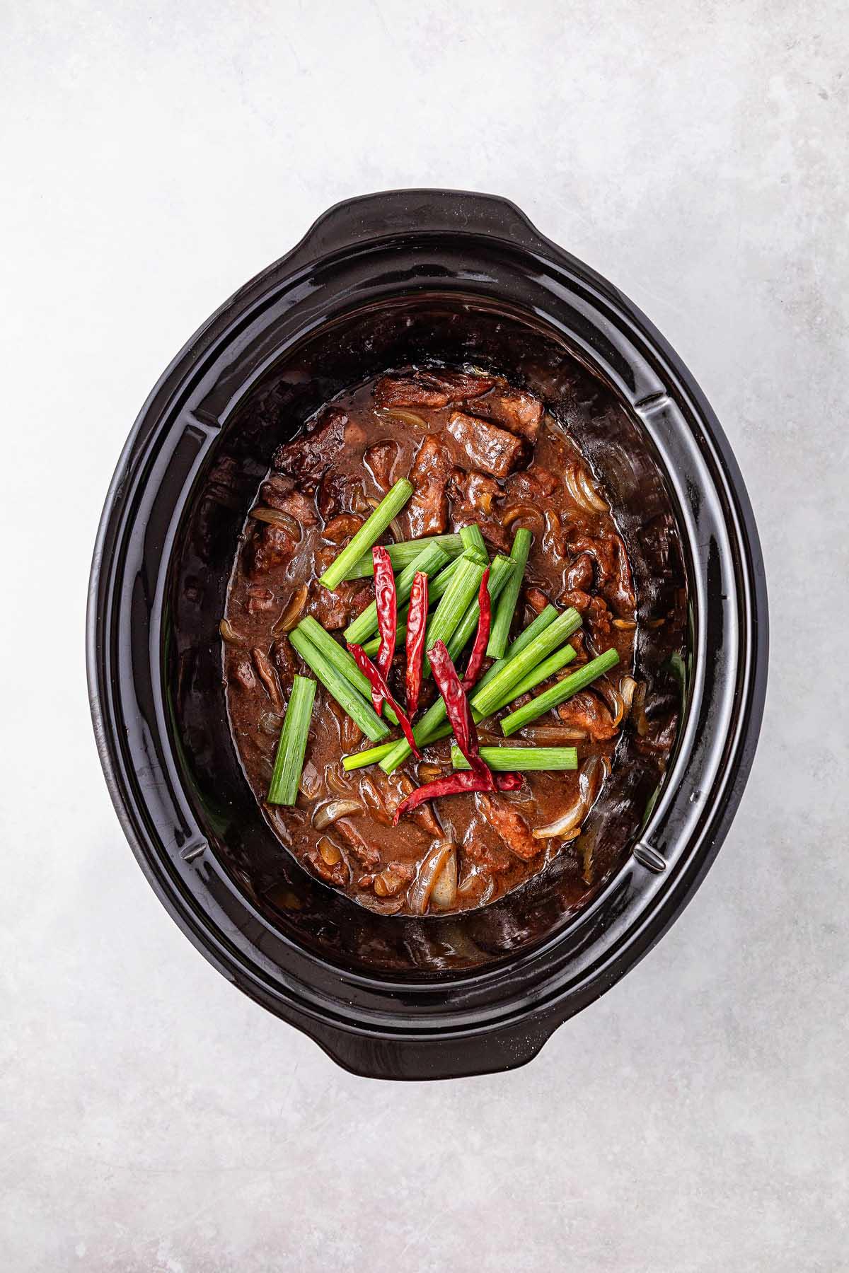 Slow cooker Mongolian beef stew topped with green onions and red chili peppers, elegantly presented on a pristine white surface.