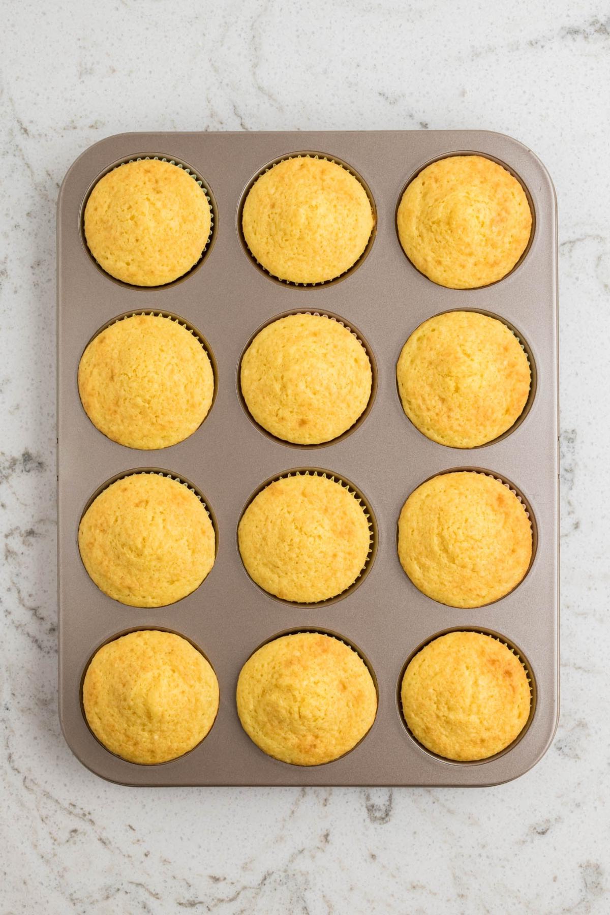 Ένα ταψί για muffins με δώδεκα φρεσκοψημένα, χρυσαφένια muffins καλαμποκιού σε μαρμάρινο πάγκο.