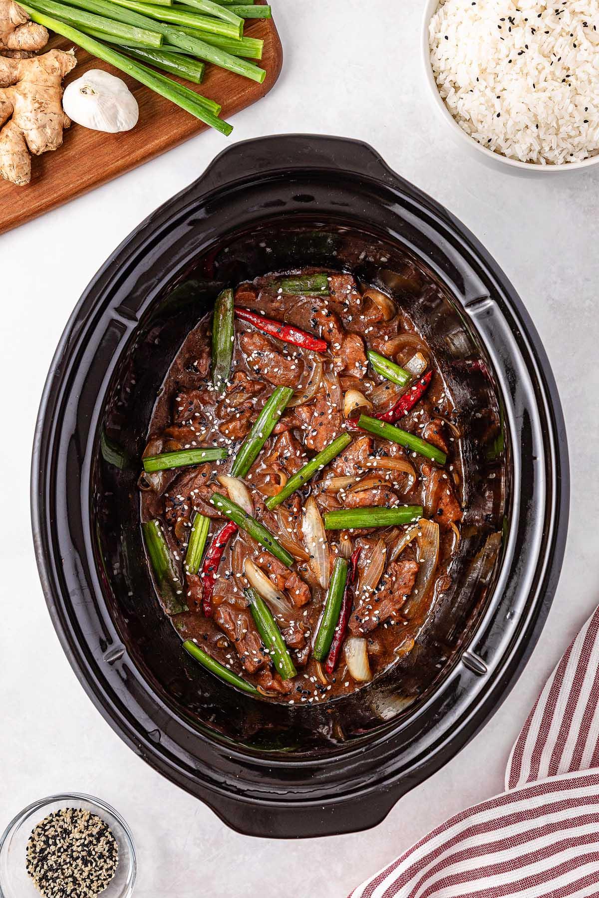 The slow cooker Mongolian beef is packed with stir-fried beef strips, green onions, and red chili peppers. Rice, ginger, and black sesame seeds are arranged beside the cooker, while a striped cloth peeks into view.