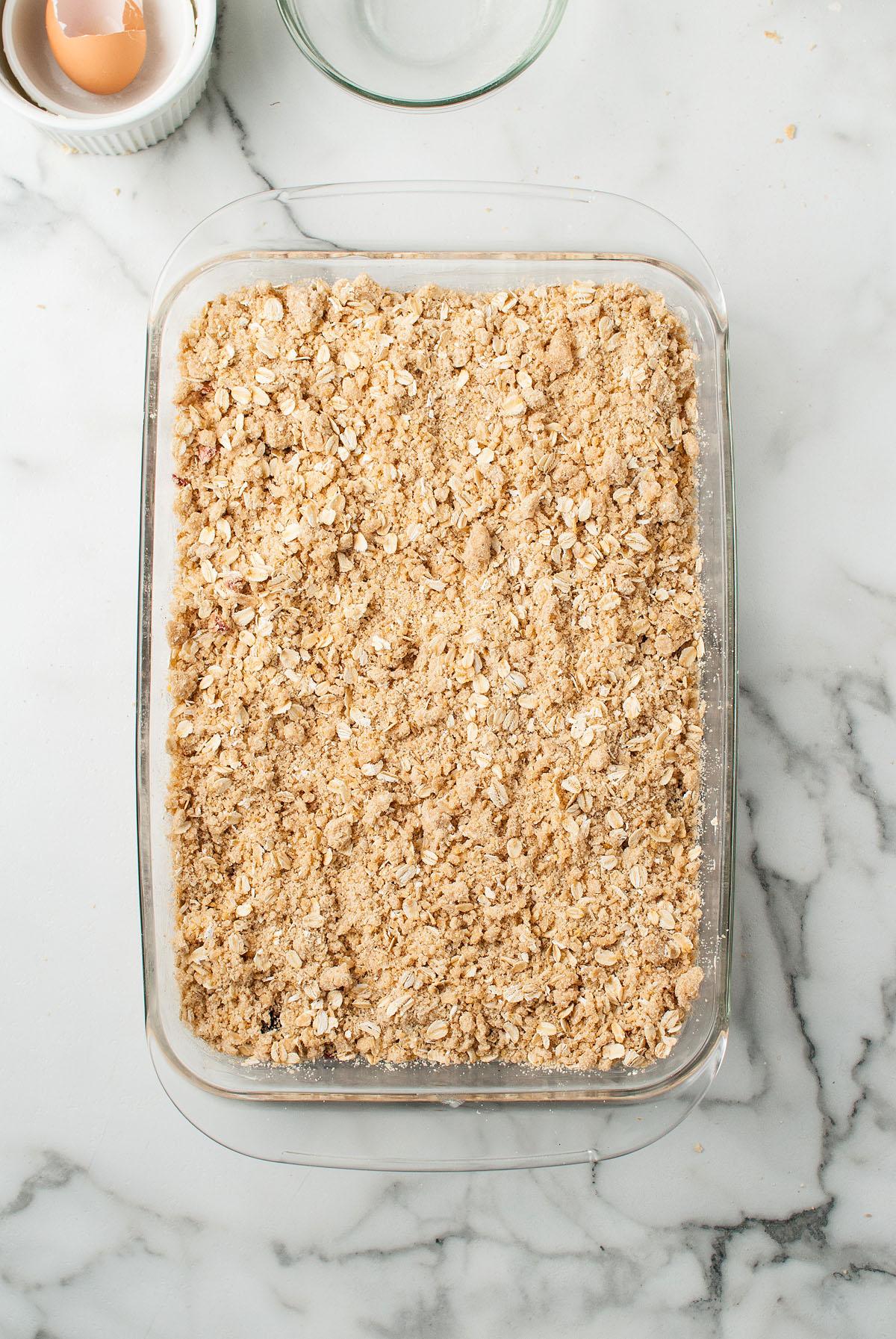Ένα γυάλινο ταψί γεμάτο με επικάλυψη crumble βρώμης για μπάρες βρώμης με γέμιση κερασιού βρίσκεται σε ένα μαρμάρινο πάγκο, δίπλα σε ένα άδειο τσόφλι αυγού.