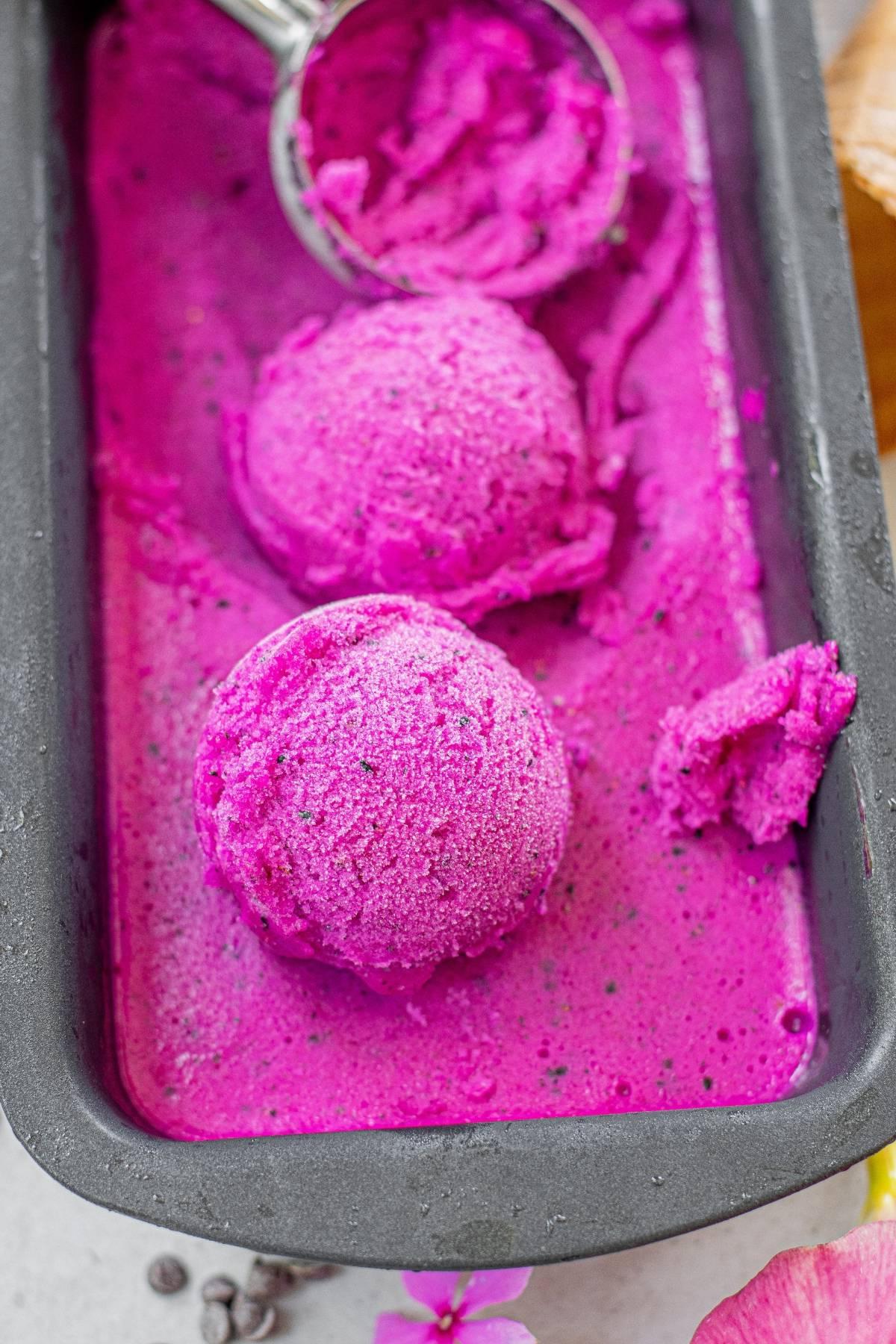 Ζωηρές ροζ μπάλες παγωτού Dragon Fruit σε ένα μαύρο ταψί με μια κουτάλα παγωτού—ένα νόστιμο παγωμένο επιδόρπιο χωρίς γαλακτοκομικά που είναι τόσο όμορφο όσο και νόστιμο.
