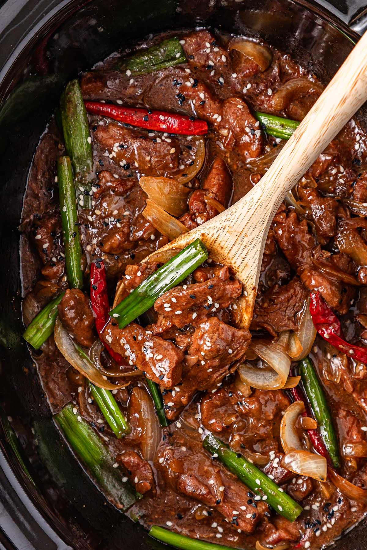 A wooden spoon stirs a slow cooker Mongolian beef dish with onions, green onions, red chilies, and sesame seeds in a rich brown sauce.