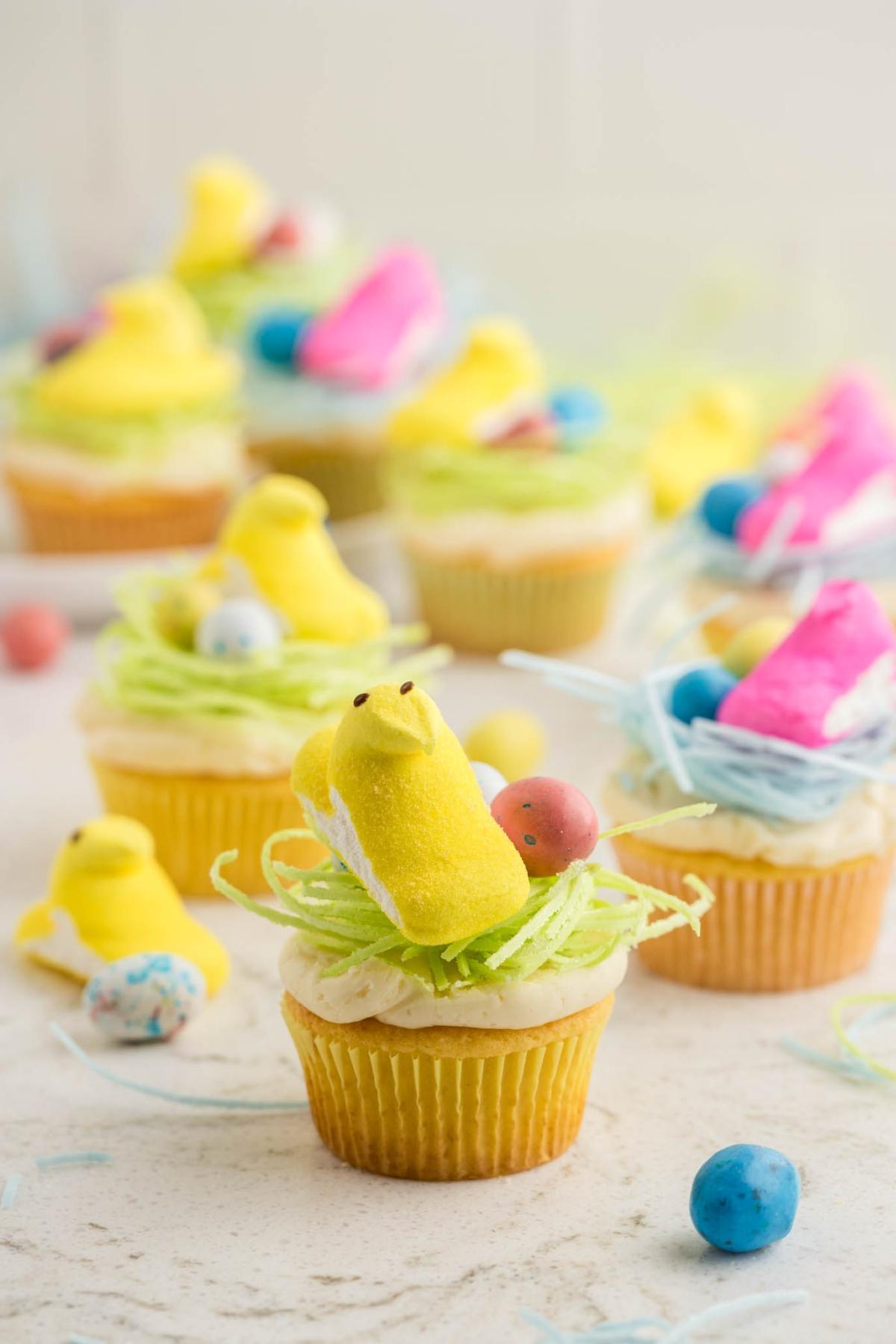 Cupcakes στολισμένα με κίτρινα μαρσμέλοου κοτόπουλα, φωλιές από πράσινη τριμμένη καρύδα και πολύχρωμα ζαχαρωτά αυγά. Άλλα διακοσμημένα cupcakes και ζαχαρωτά είναι θολά στο παρασκήνιο.
