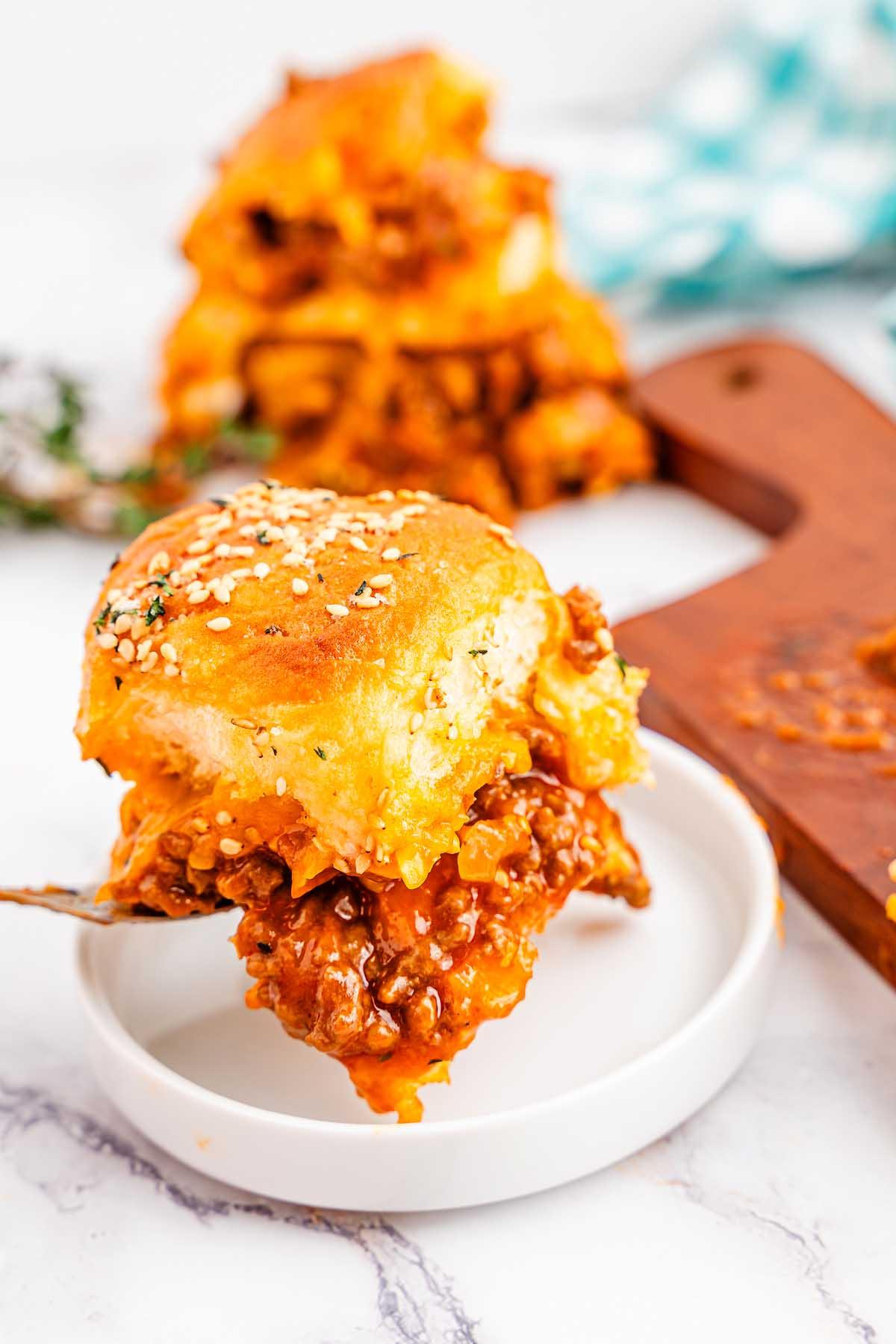 Κοντινό πλάνο ενός τυρένιου slider Sloppy Joe με σουσάμι, γεμιστό με κιμά και σάλτσα, σε ένα μικρό λευκό πιάτο. Περισσότερα sliders είναι ορατά στο φόντο σε μια ξύλινη επιφάνεια κοπής.