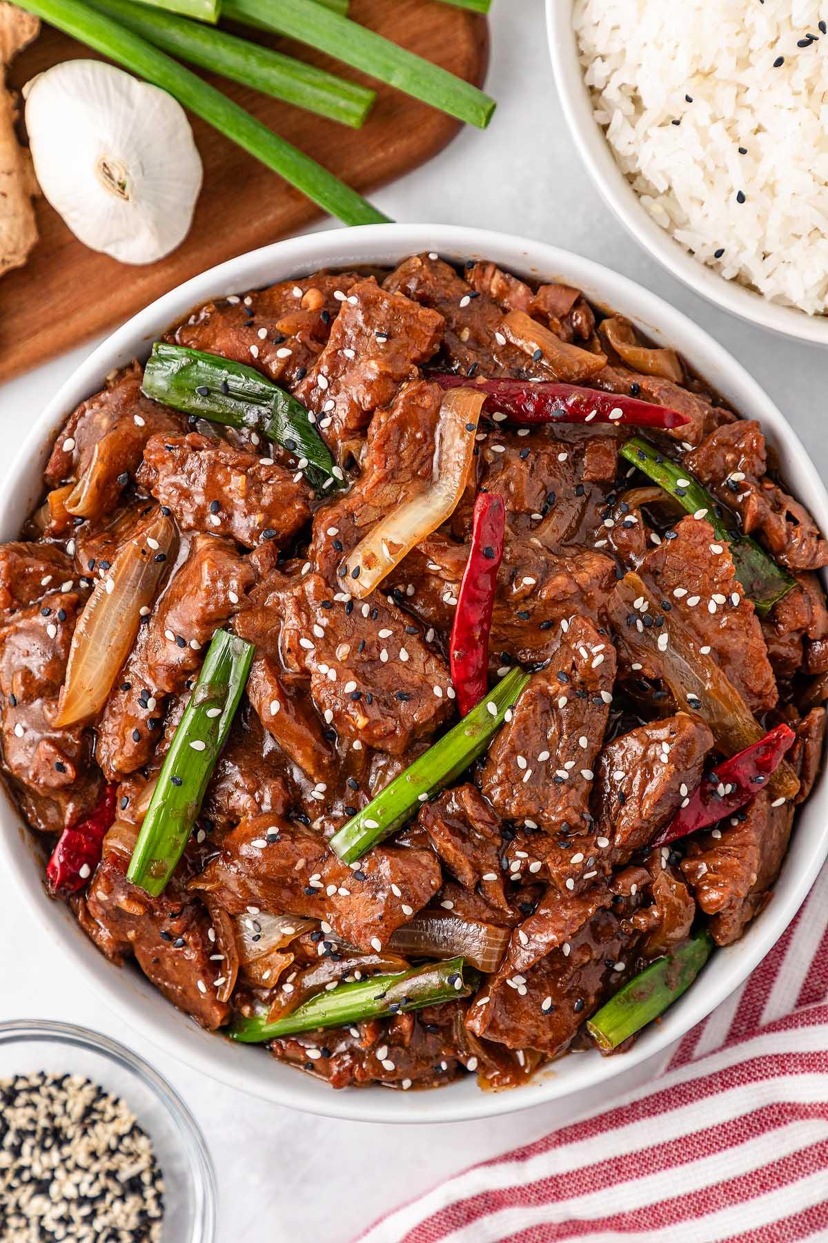 A bowl of stir-fried beef with green onions and red chilies, garnished with sesame seeds, reminiscent of slow cooker Mongolian beef. Rice, sliced green onions, garlic, and ginger sit invitingly in the background.