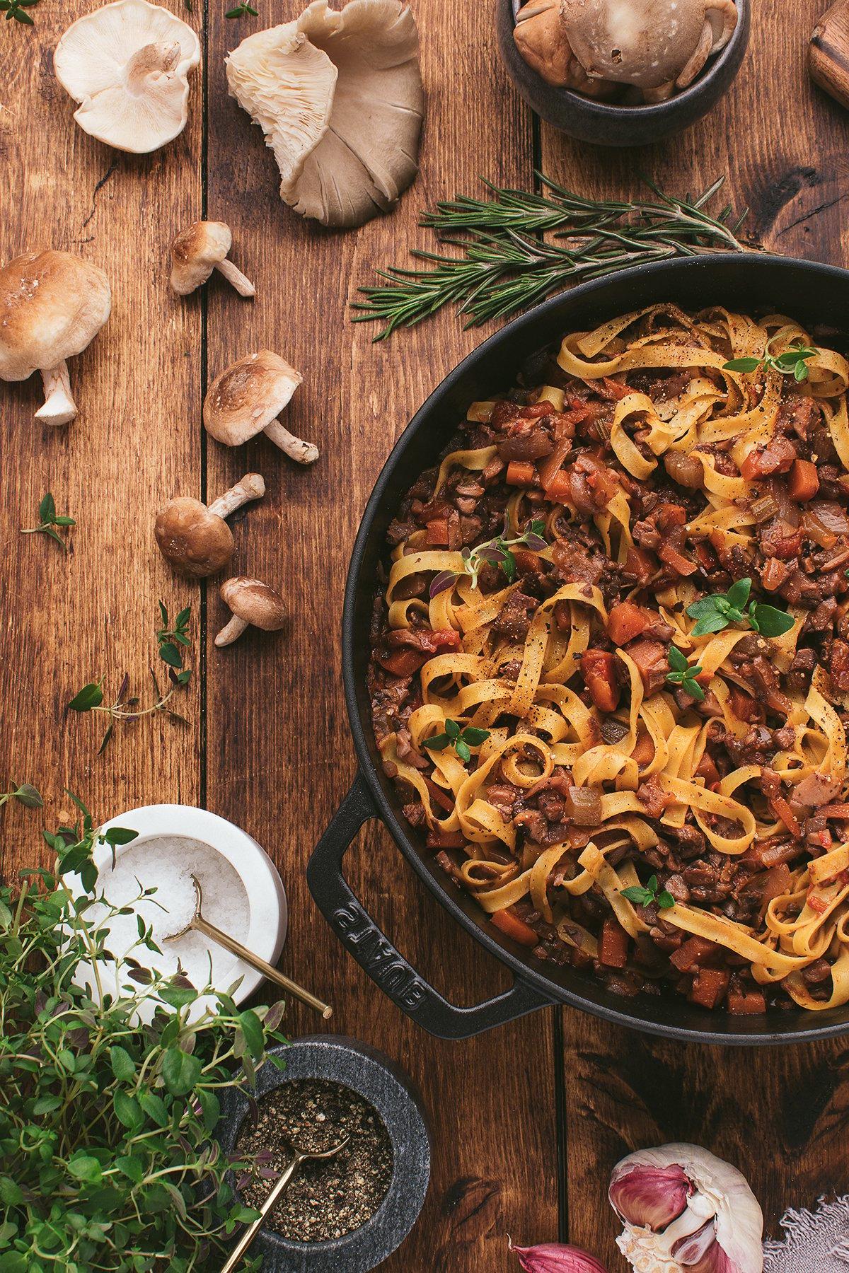 Τα tagliatelle ζυμαρικά με κρεμώδη βίγκαν σάλτσα ζυμαρικών και μανιτάρια σε ένα τηγάνι, περιτριγυρισμένα από φρέσκα βότανα και ολόκληρα μανιτάρια πάνω σε ένα ξύλινο τραπέζι.