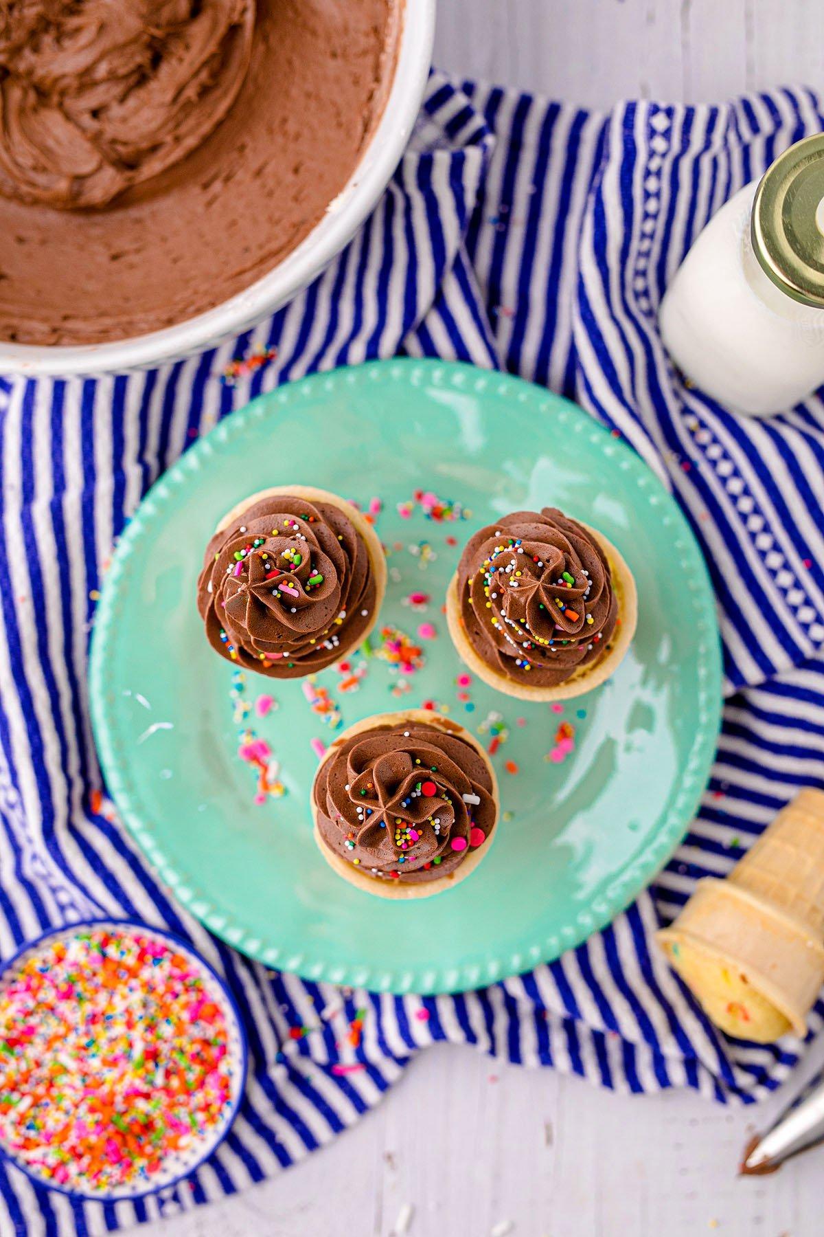 Τρία cupcakes με σοκολατένια βουτυρόκρεμα και τρούφα σε ένα πράσινο πιάτο, περιτριγυρισμένα από συστατικά ψησίματος – τέλεια cupcakes σε χωνάκι παγωτού για πάρτι γενεθλίων.