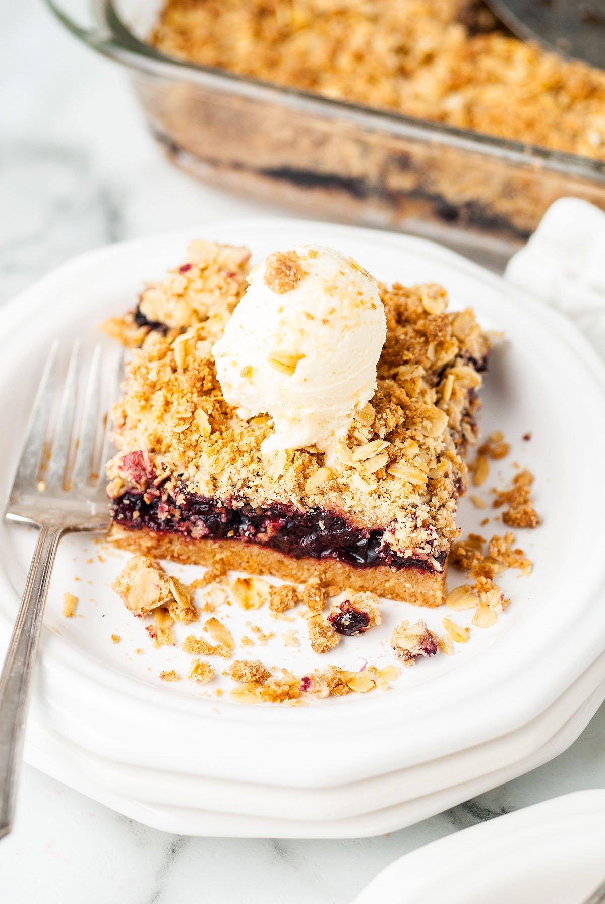 Μια φέτα από crumble με μούρα, εμπνευσμένη από μια συνταγή copycat της Starbucks, με μια μπάλα παγωτό βανίλια σε ένα λευκό πιάτο με ένα πιρούνι—γευστικά θυμίζει μπάρες βρώμης με γέμιση κερασιού από κατεψυγμένα κεράσια.
