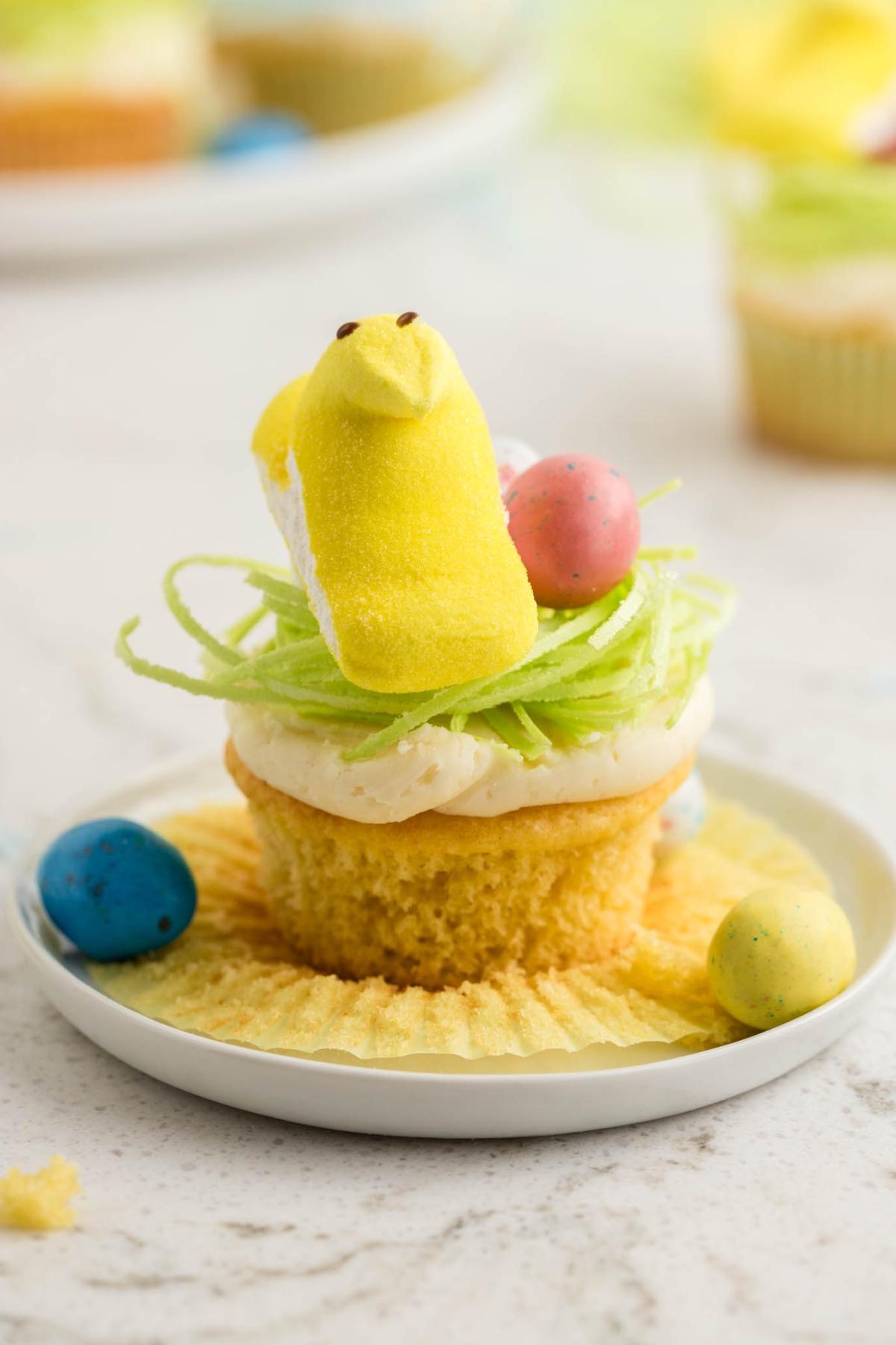 Ένα cupcake στολισμένο με ένα κίτρινο κοτόπουλο μαρσμέλοου, χρωματιστά αυγά και πράσινες διακοσμήσεις, τοποθετημένο σε ένα λευκό πιάτο.