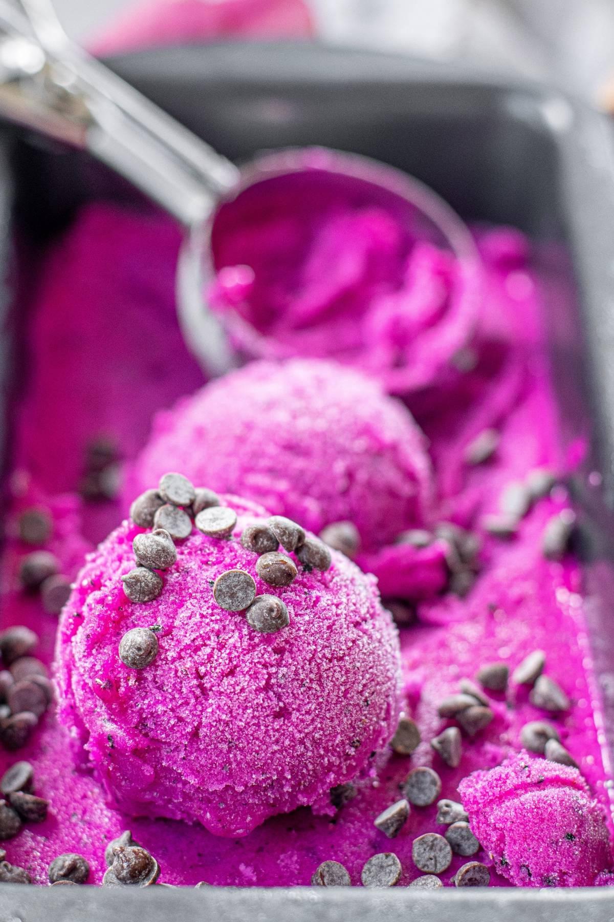 Δύο μπάλες φωτεινού ροζ παγωτού dragon fruit με κομματάκια σοκολάτας από πάνω, σε ένα δίσκο με μεταλλική σπάτουλα παγωτού—ένα ζωντανό και πολύχρωμο γευστικό παγωτό που είναι τόσο νόστιμο όσο φαίνεται.