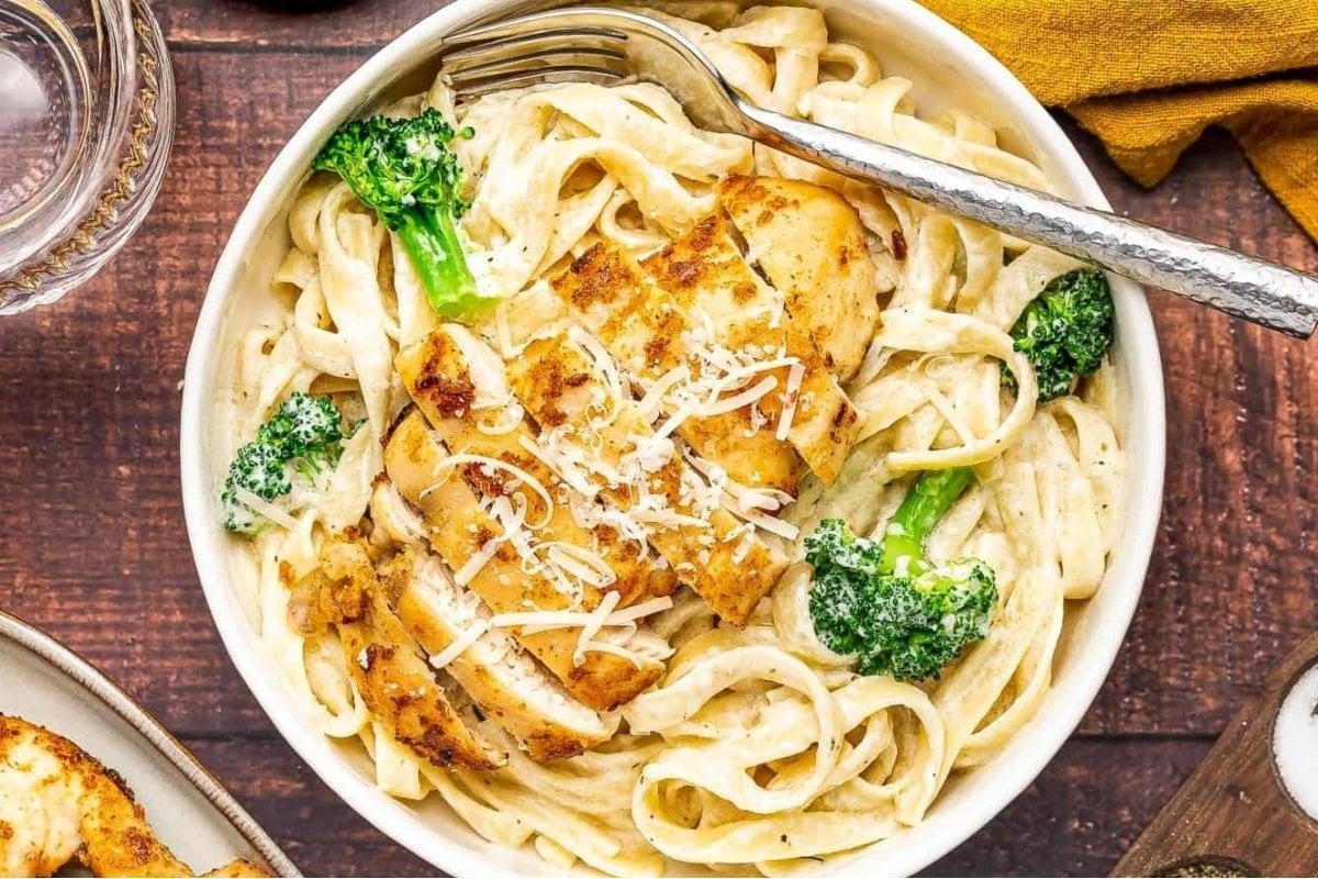 Ένα μπολ με fettuccine alfredo με μπρόκολο και ψητό κοτόπουλο, γαρνιρισμένο με τριμμένη παρμεζάνα. Ένα πιρούνι ακουμπά στο χείλος του μπολ, καθιστώντας το ένα τέλειο πιάτο για όσους εξερευνούν νόστιμες συνταγές fettuccine.