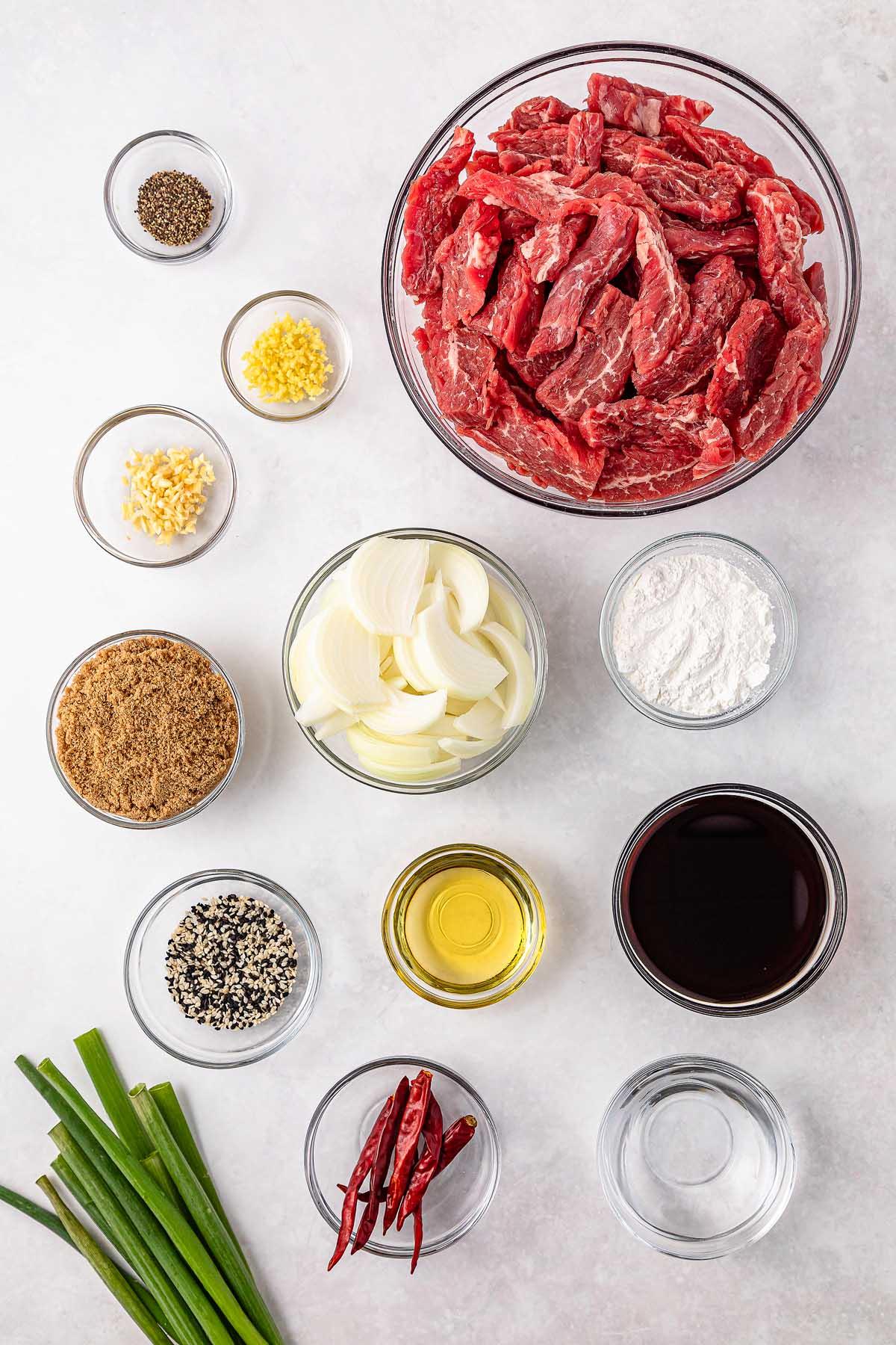 Ingredients for slow cooker Mongolian beef are thoughtfully arranged: sliced beef, onions, minced garlic and ginger, ground black pepper, soy sauce, brown sugar, sesame seeds, chili peppers, cooking oil, flour, and green onions.