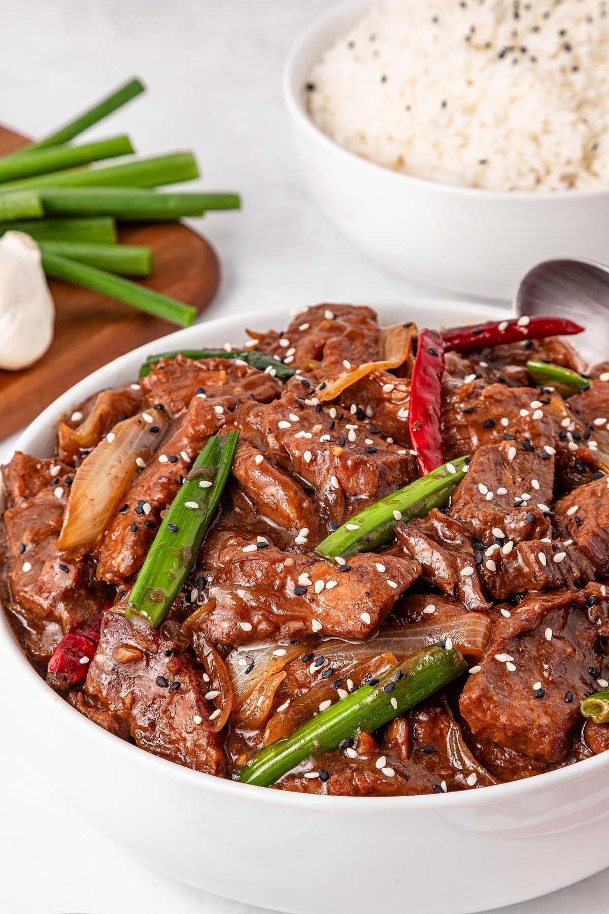 A bowl of Mongolian beef stir-fry garnished with green onions, sesame seeds, and red chilies. In the background, a bowl of white rice sits alongside fresh green onions, rounding out this Asian-inspired feast.