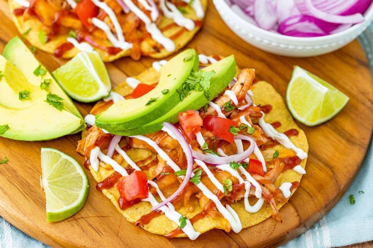 Δύο tostadas με τεμαχισμένο κοτόπουλο, κομμένο αβοκάντο, κρεμμύδια, ντομάτα, και περιχυμένα με σάλτσα, σερβιρισμένα με φέτες λάιμ σε ξύλινη σανίδα. Ιδανικό για όσους αναζητούν νόστιμες συνταγές κοτόπουλου.