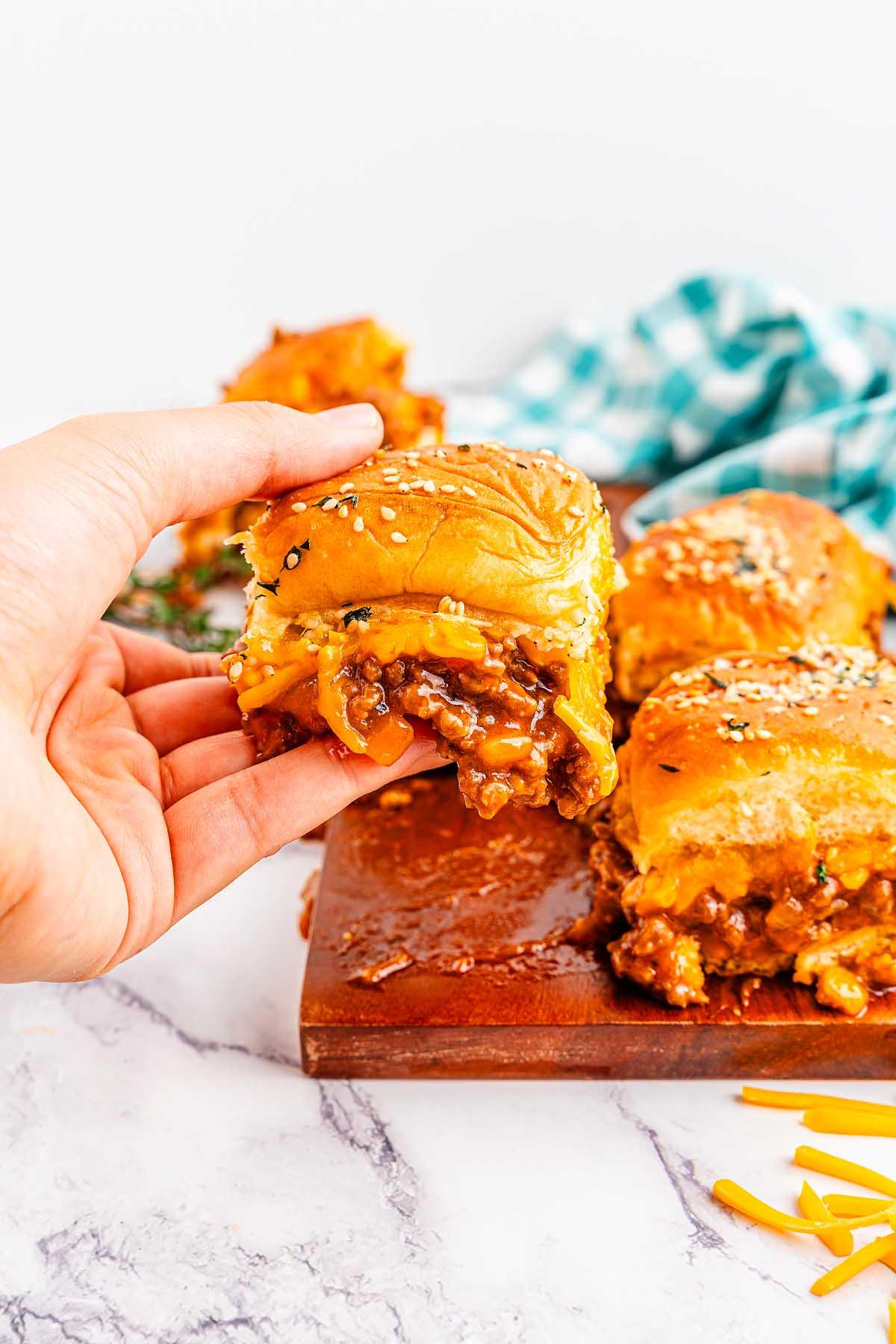 Χέρι που κρατά ένα σάντουιτς Sloppy Joe. Κομμάτια τριμμένου τυριού βρίσκονται στο φόντο σε ξύλινη επιφάνεια και μαρμάρινη επιφάνεια.