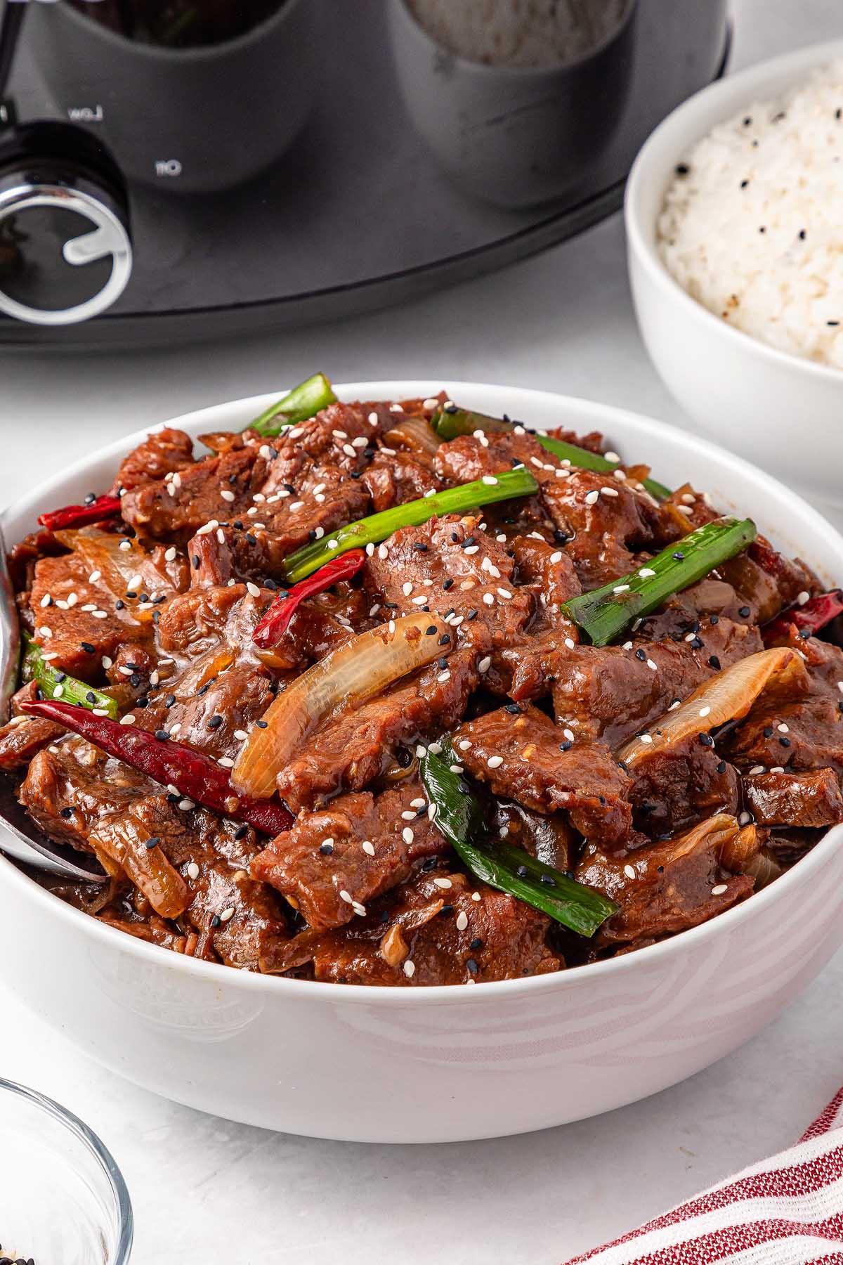 A bowl of slow cooker Mongolian beef with green onions and red chilies, sprinkled with sesame seeds. A bowl of rice sits in the background.