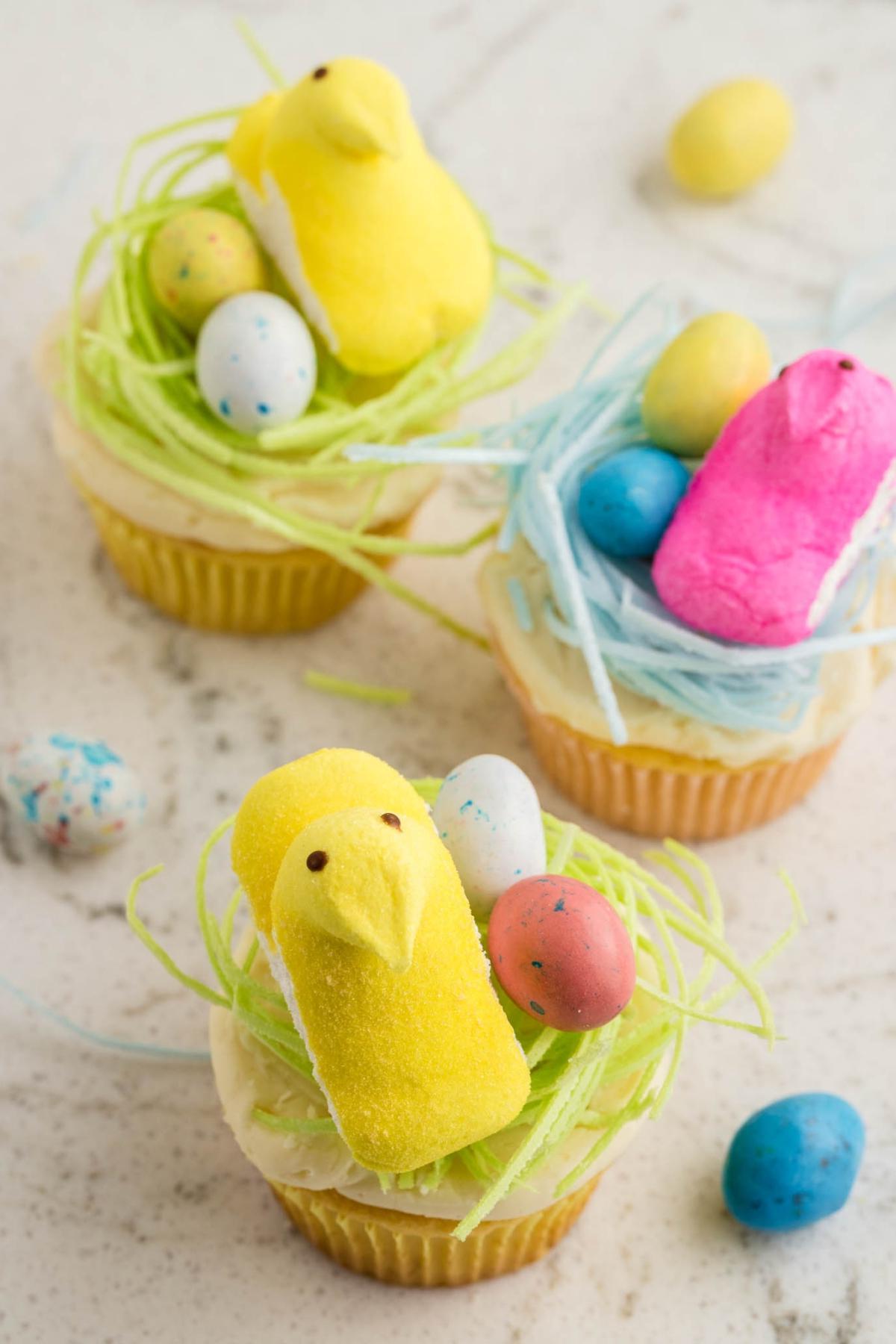 Cupcakes διακοσμημένα με πράσινο και μπλε γλάσο, στολισμένα με πολύχρωμα μαρσμέλοου κοτόπουλα και στικτά αυγά σε μαρμάρινη επιφάνεια.
