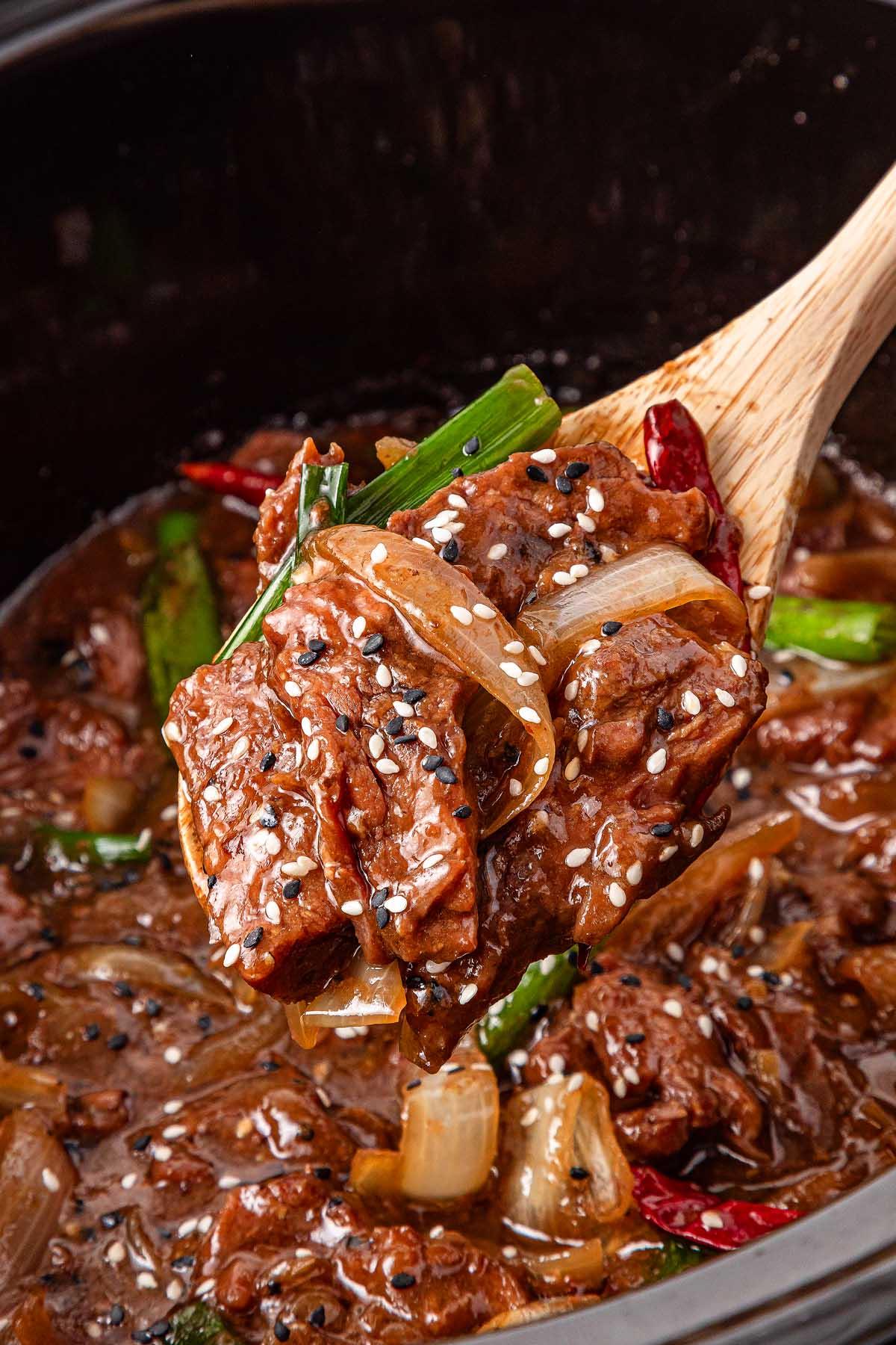 A wooden spoon cradles a portion of slow cooker Mongolian beef stew with onions, green peppers, and sesame seeds. The dish looks rich and saucy, while a crockpot brimming with more savory stew waits in the background.