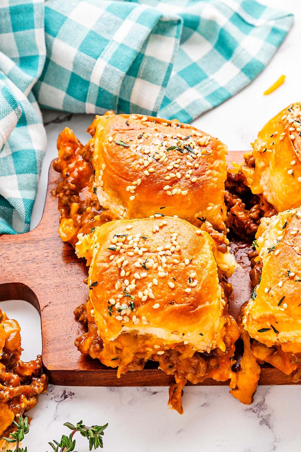 Sliders με ψωμάκια με σουσάμι γεμισμένα με ένα αλμυρό μείγμα Sloppy Joe και τυρί, τοποθετημένα σε ξύλινη σανίδα δίπλα σε ένα μπλε και λευκό καρό ύφασμα.