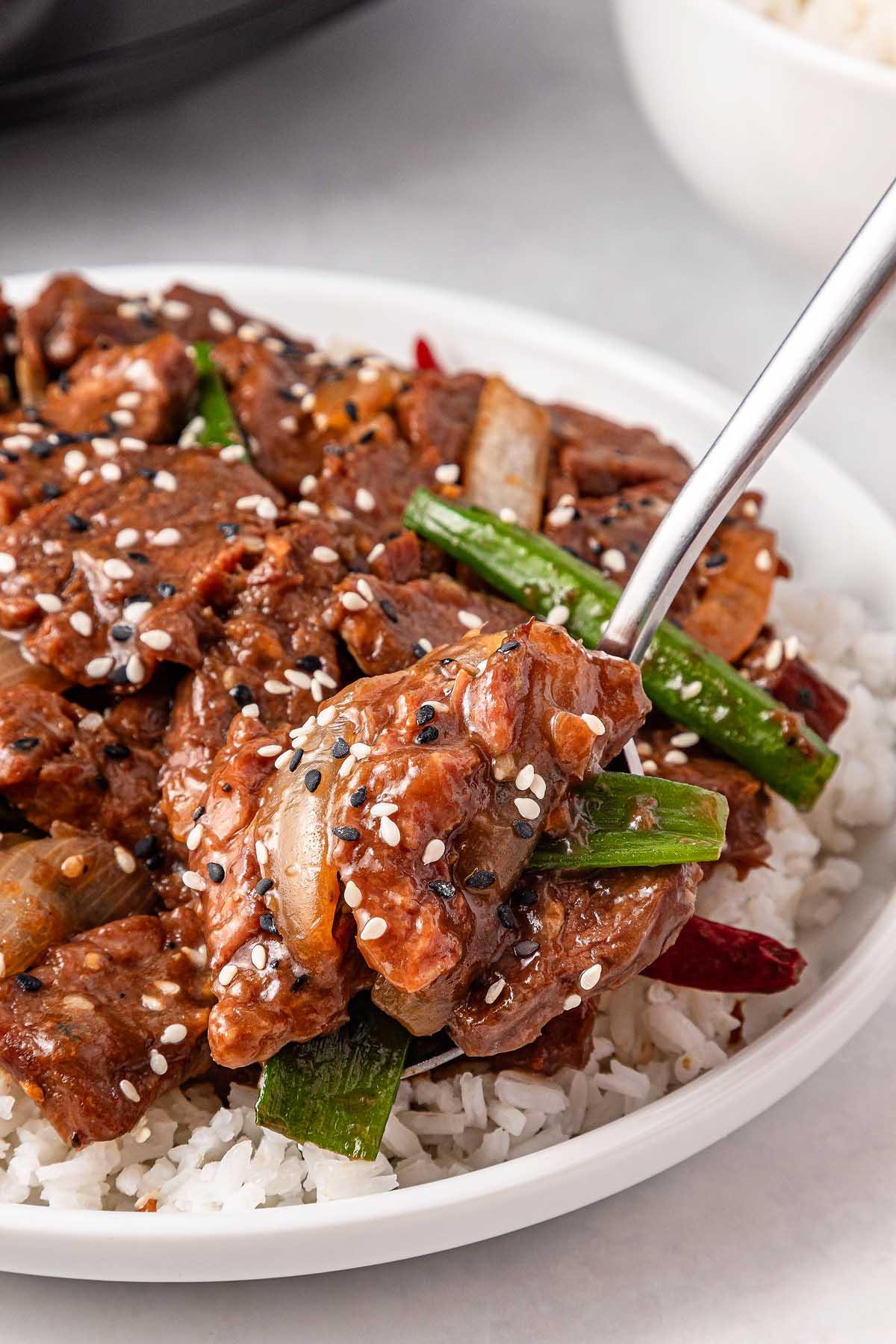A plate of stir-fry featuring tender slow cooker Mongolian beef, green peppers, onions, and red chili peppers on rice, garnished with black and white sesame seeds.