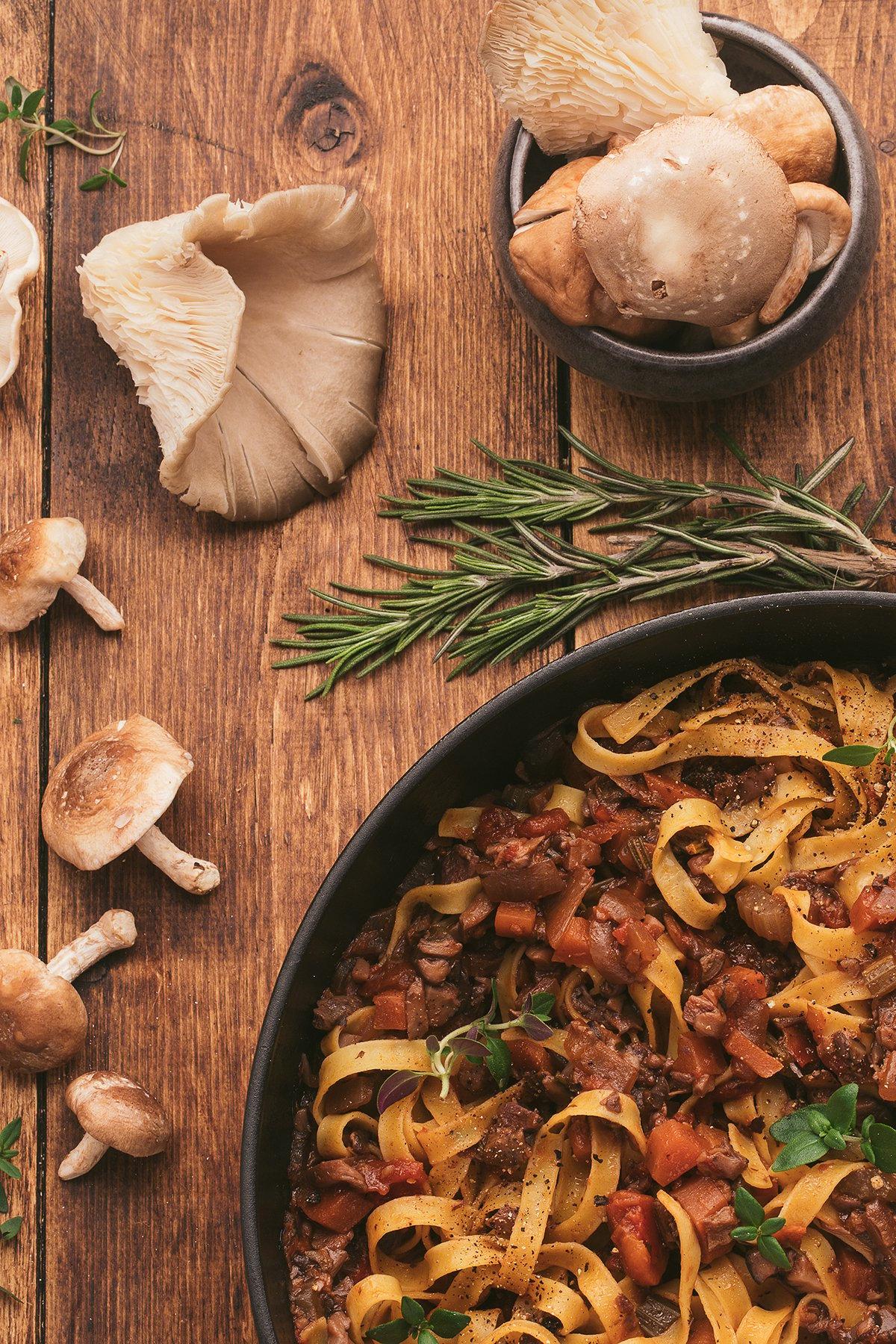 Τα tagliatelle ζυμαρικά με πλούσια βίγκαν σάλτσα ζυμαρικών σε ένα τηγάνι, περιτριγυρισμένα από μανιτάρια και φρέσκο δεντρολίβανο πάνω σε ένα ξύλινο τραπέζι.