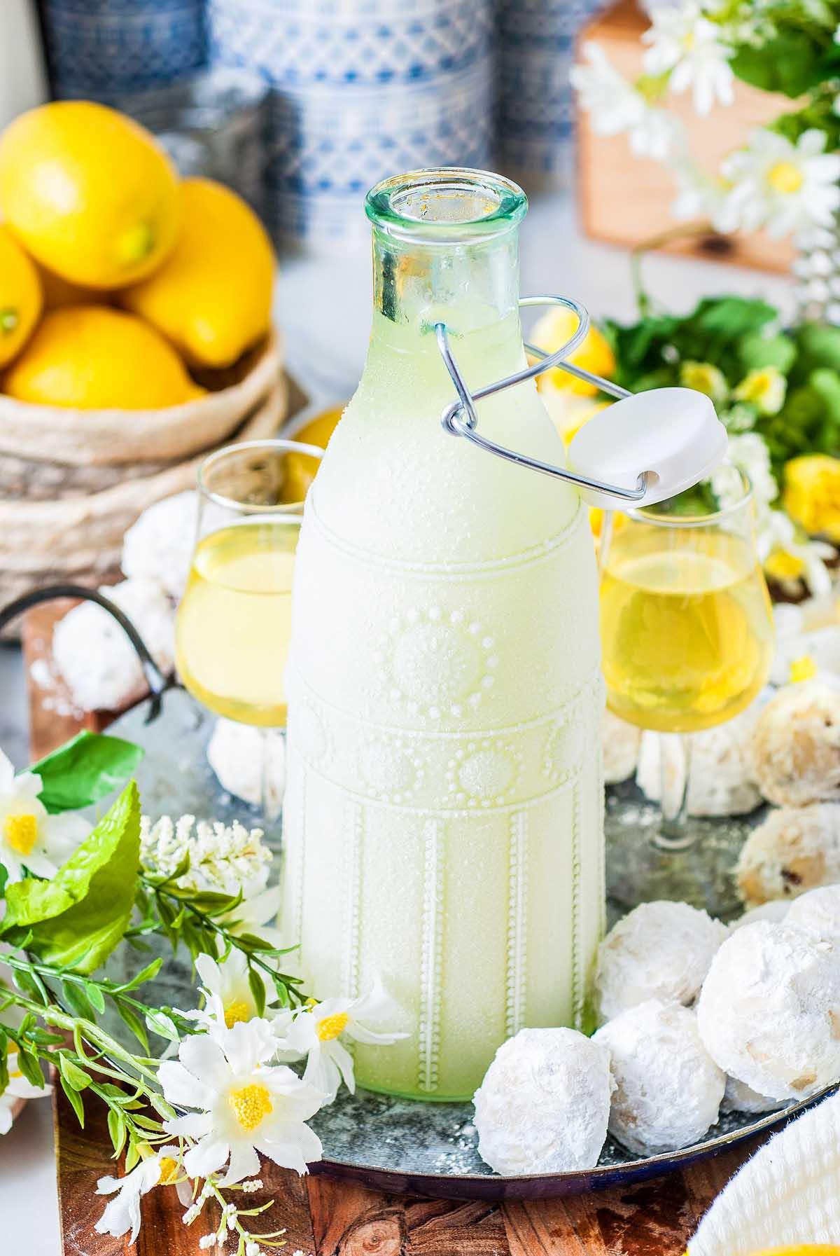 Ένα γυάλινο μπουκάλι limoncello, φτιαγμένο από μια παραδοσιακή ιταλική συνταγή limoncello, στέκεται κομψά με δύο ποτήρια, περιτριγυρισμένο από ζαχαρωμένα αρτοσκευάσματα, ζωντανά λουλούδια και φρέσκα λεμόνια σε έναν δίσκο.