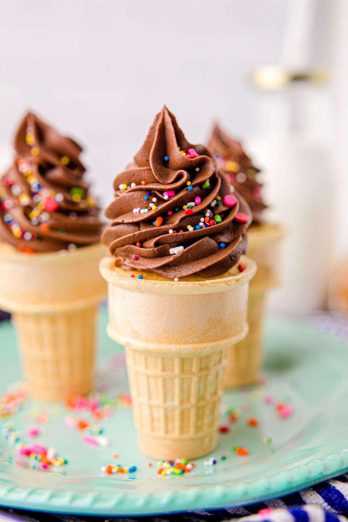 Cupcakes σε χωνάκι παγωτού με σοκολατένια βουτυρόκρεμα και πολύχρωμη τρούφα αποτελούν μια διασκεδαστική λιχουδιά, ιδανικά ως cupcakes για πάρτι γενεθλίων, σερβιρισμένα σε ένα ανοιχτό πράσινο πιάτο.