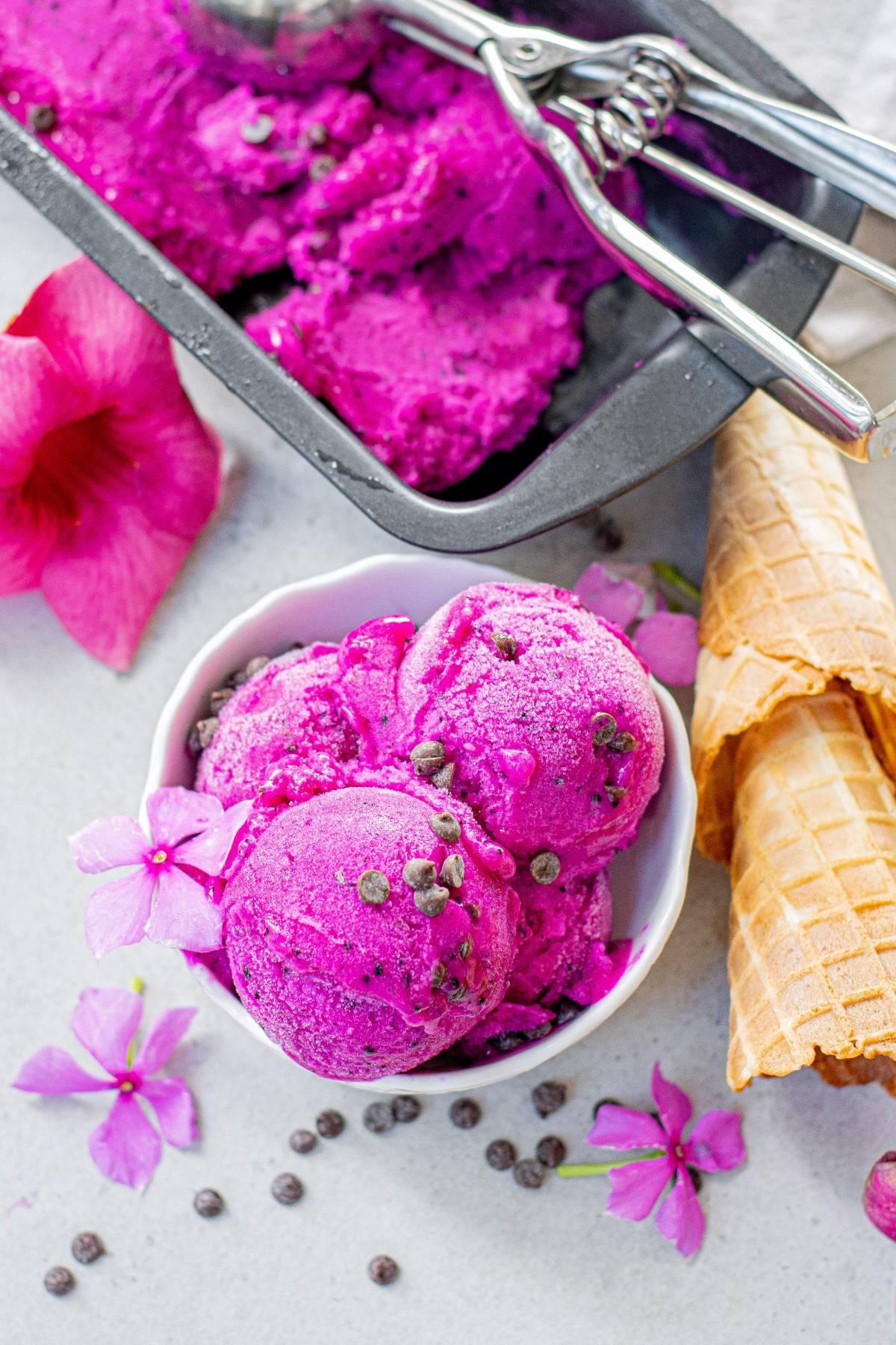 Μπολ με φωτεινό ροζ παγωτό Dragon Fruit με κομματάκια σοκολάτας, λουλούδια και χωνάκια βάφλας, δίπλα σε ένα δίσκο παγωτού—ένα ακαταμάχητα πολύχρωμο παγωτό ιδανικό για καλοκαιρινές λιχουδιές.