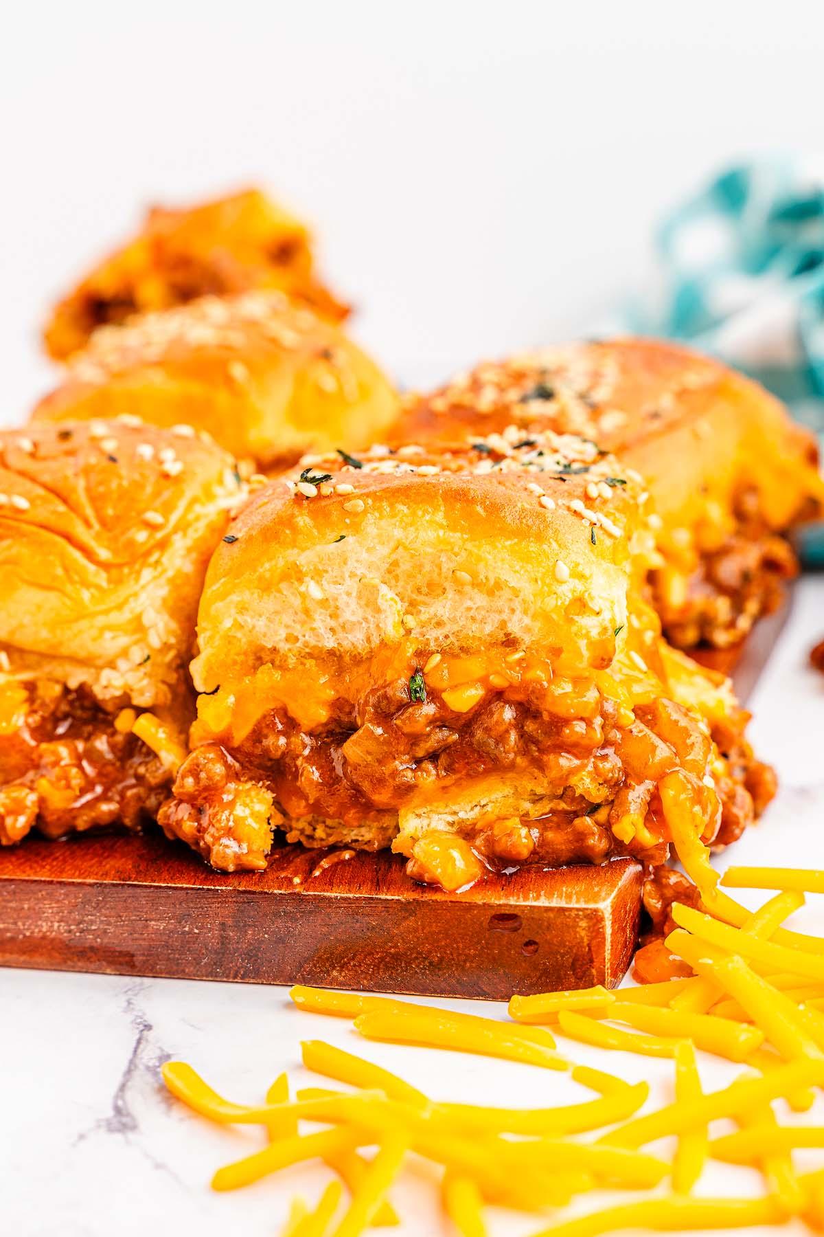 Κοντινό πλάνο τριών τυρένιων Sloppy Joe sliders με ψωμάκια με σουσάμι σε ξύλινη σανίδα. Τριμμένο τυρί είναι σκορπισμένο γύρω από τα sliders.