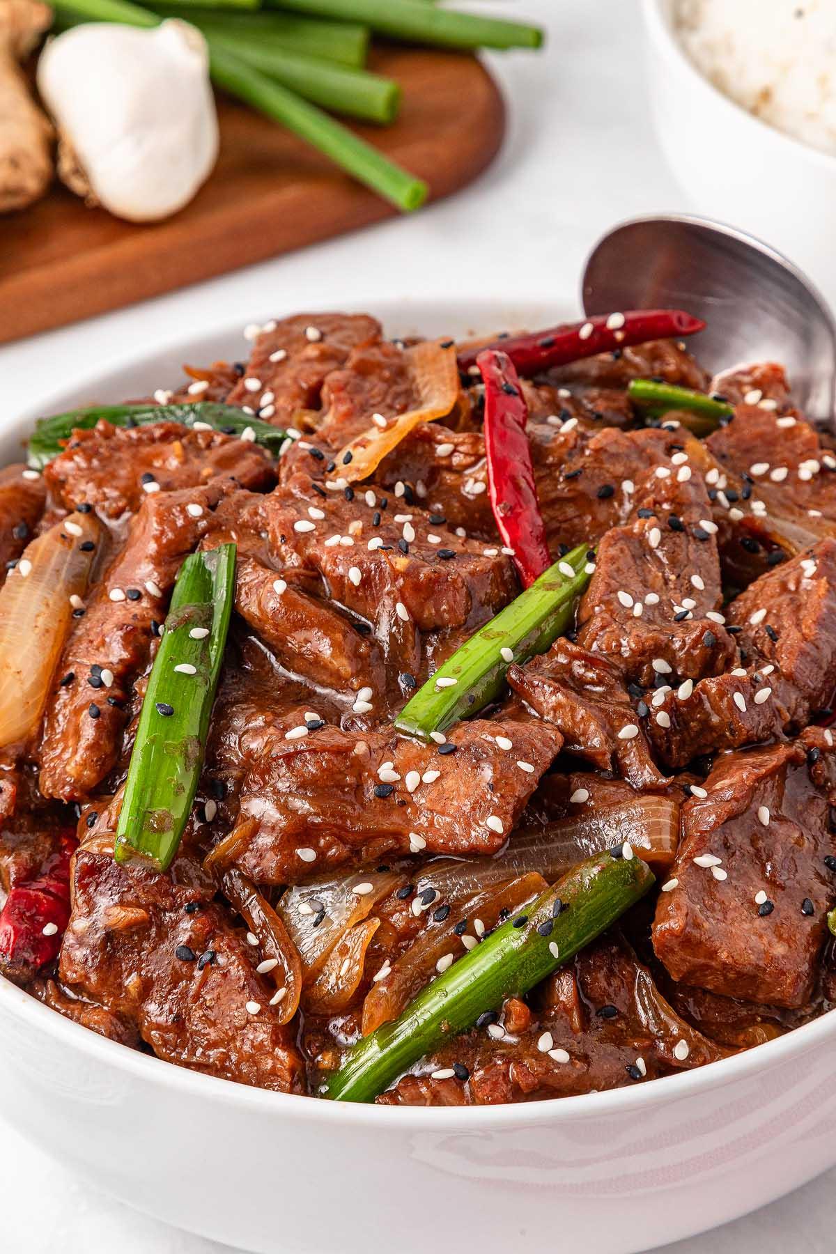A bowl of spicy slow-cooker Mongolian beef stir-fry with green onions, red chili peppers, and sesame seeds. A spoon rests inside the bowl. Garlic and more green onions are on a wooden board in the background.