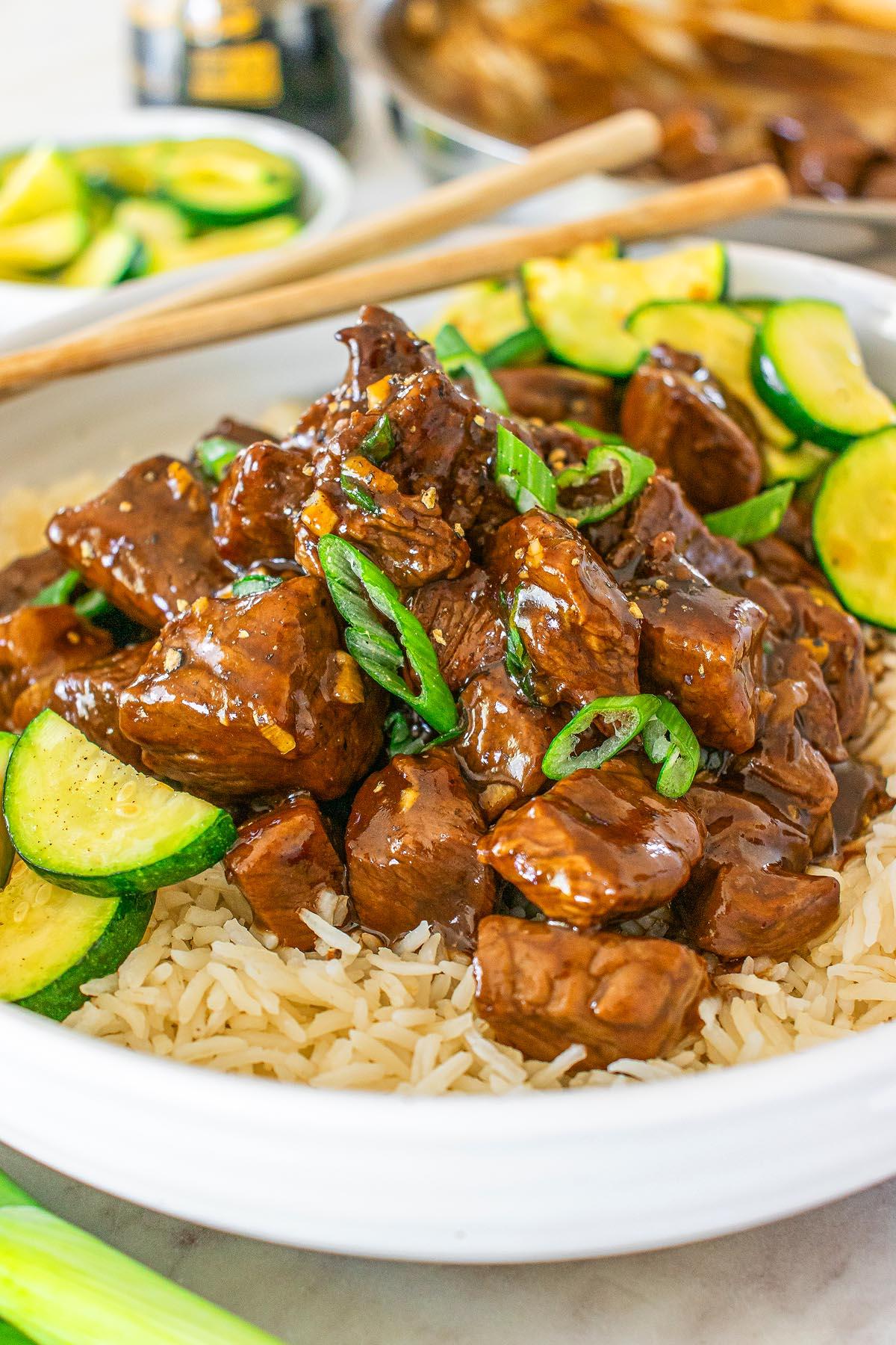 Ένα πιάτο stir-fry τεριγιάκι μοσχαριού με κολοκυθάκια σε φέτες και φρέσκα κρεμμυδάκια σερβιρισμένο πάνω σε ρύζι. Τσόπστικς ακουμπούν στην άκρη του πιάτου.