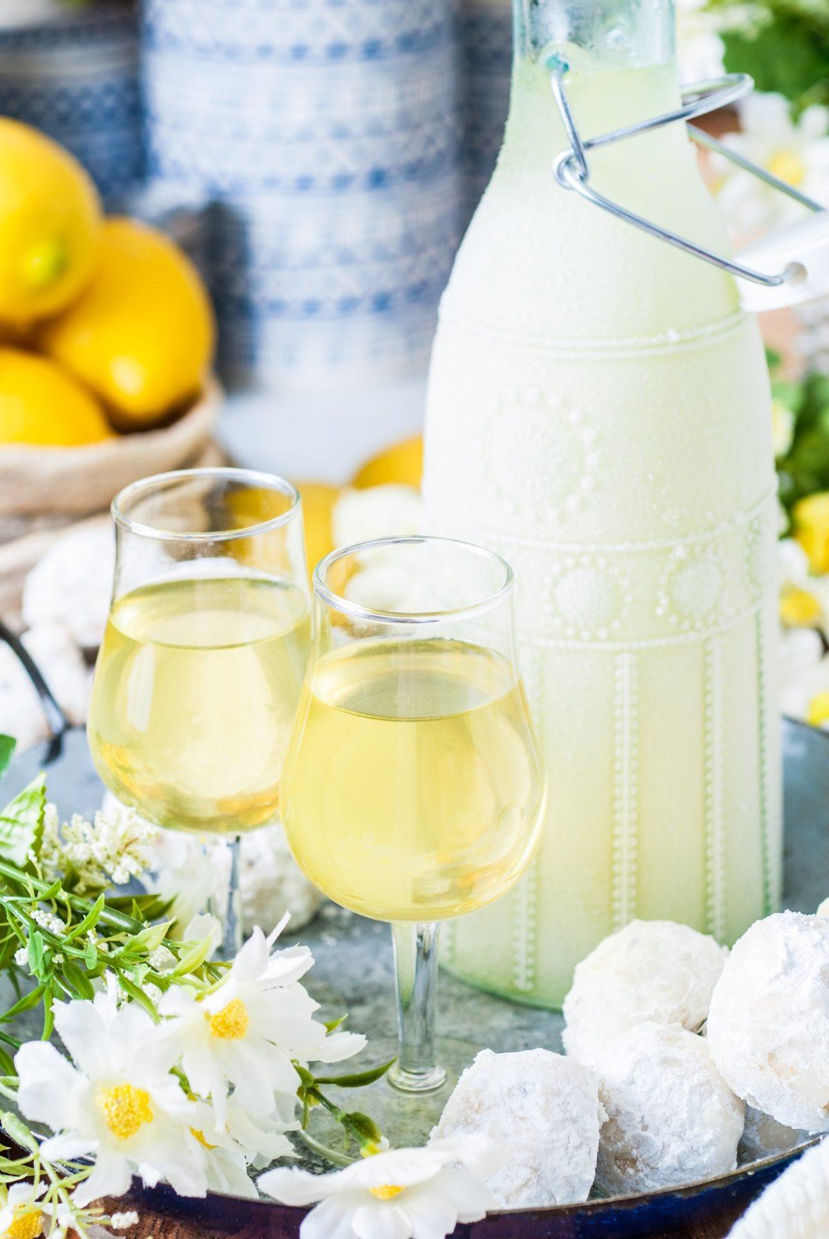 Δύο ποτήρια limoncello κοσμούν έναν δίσκο δίπλα σε ένα διακοσμητικό μπουκάλι και λευκά λουλούδια, αναδεικνύοντας μια ιταλική συνταγή limoncello. Στο φόντο, λεμόνια και μπουκάλια με σχέδια προσθέτουν μια νότα κομψότητας.