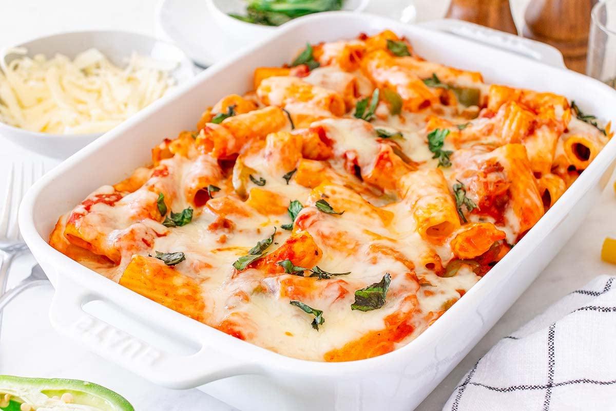 Ένα πιάτο Pasta al Forno με λιωμένο τυρί και βότανα σε ένα λευκό πυρίμαχο σκεύος, με τριμμένο τυρί στο παρασκήνιο.