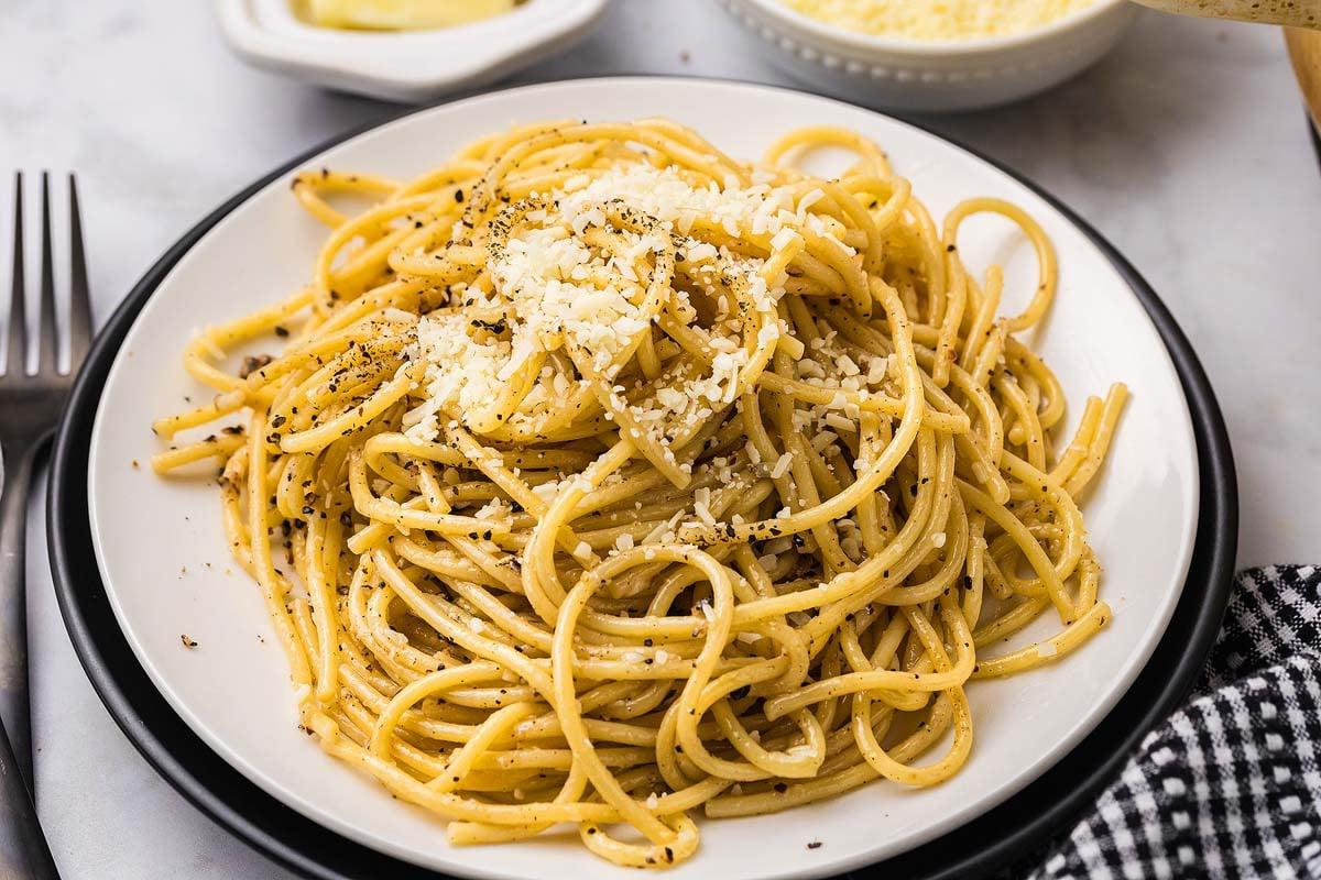 Ένα αχνιστό πιάτο cacio e pepe, στριμμένο με τριμμένο τυρί και μαύρο πιπέρι, βρίσκεται δελεαστικά στο τραπέζι, συνοδευόμενο από μια καρό πετσέτα και ένα γυαλιστερό πιρούνι.