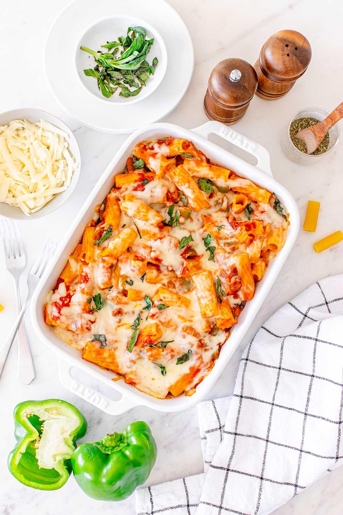 Ένα ταψί τυρένιο Pasta al Forno σε ένα τραπέζι με μισές πιπεριές, τριμμένο τυρί, μια ασπρόμαυρη καρό πετσέτα, αλατοπιπεριέρες και ένα μικρό μπολ με βότανα.