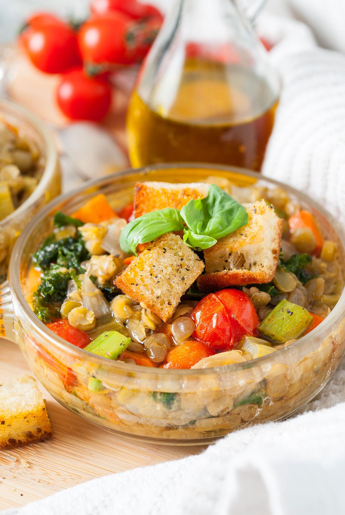 Ένα μπολ με ιταλική σούπα φακής – Zuppa Di Lenticchie με φακές, κρουτόν και φρέσκο βασιλικό, με ελαιόλαδο και ντομάτες στο φόντο.