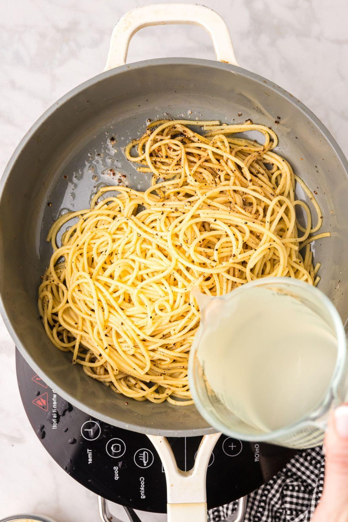 Ένα άτομο ετοιμάζει επιδέξια cacio e pepe χύνοντας υγρό από μια μεζούρα σε ένα τηγάνι με σπαγγέτι.